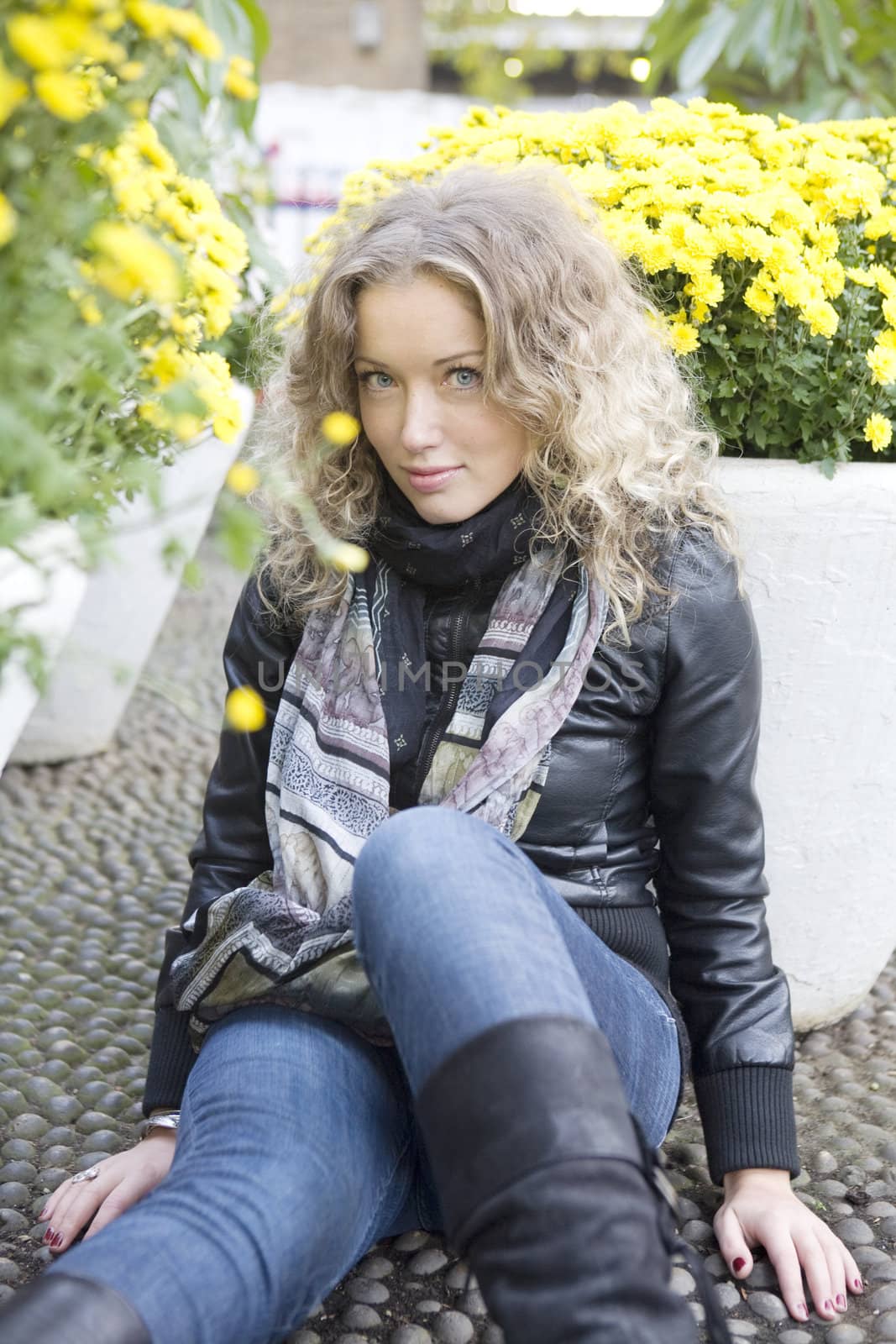 portrait of young attractive blond girl with curl hair sitting on the stone-block pavement between two vase of flower in the park
