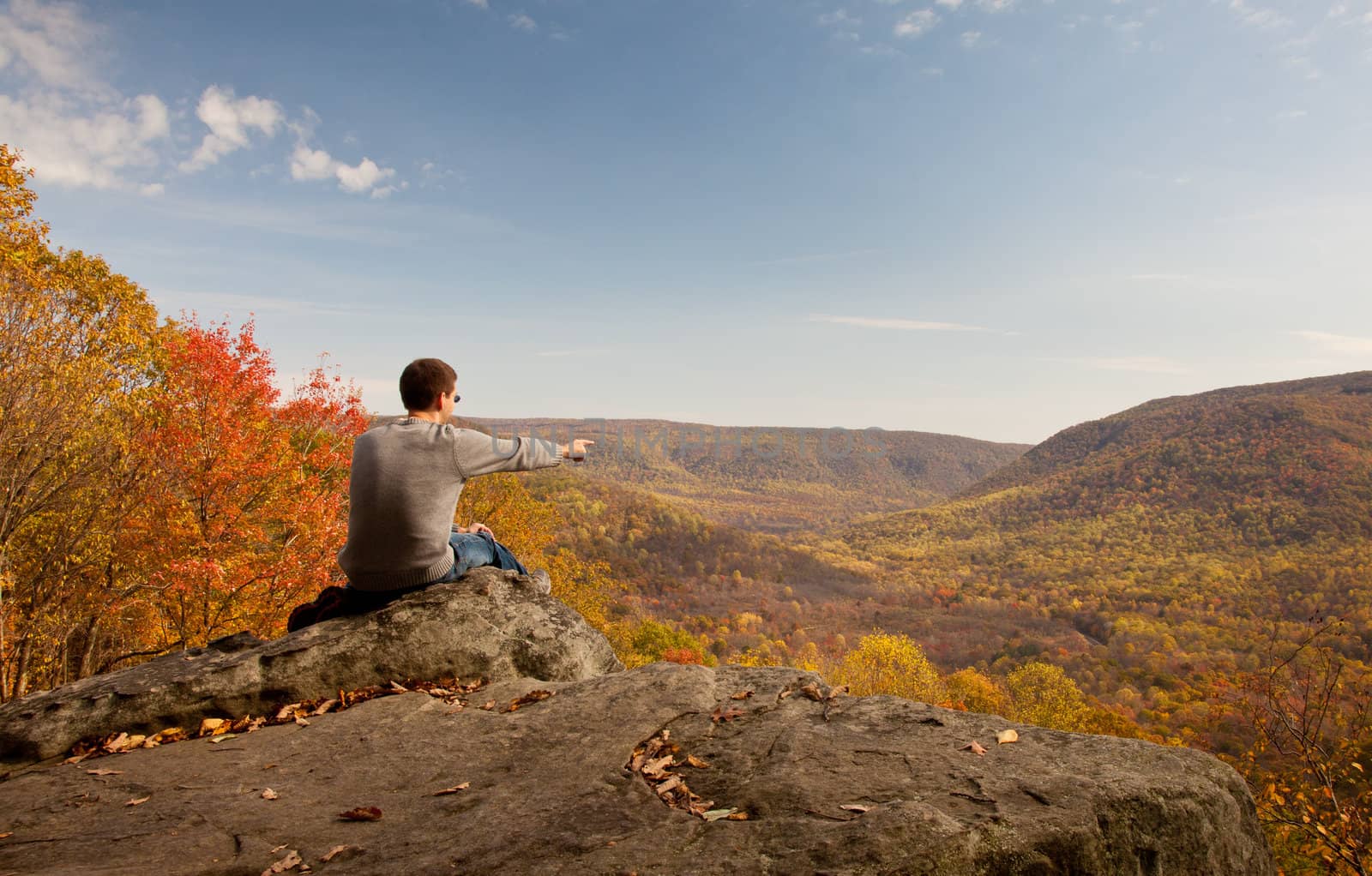 Young male hiker in jeans pondering while sitting on a boulder in front of fall leaves