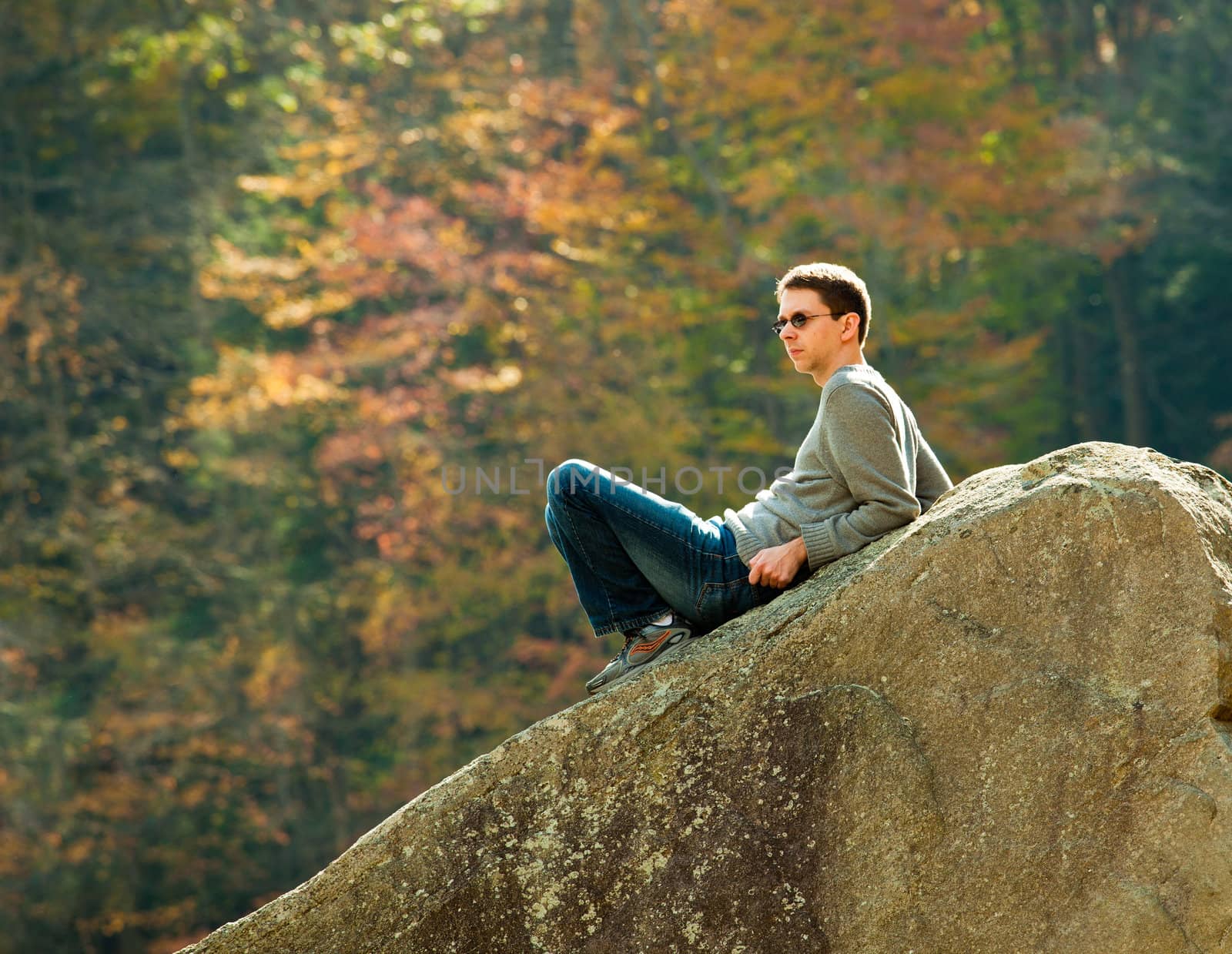 Young male hiker in jeans pondering while sitting on a boulder in front of fall leaves