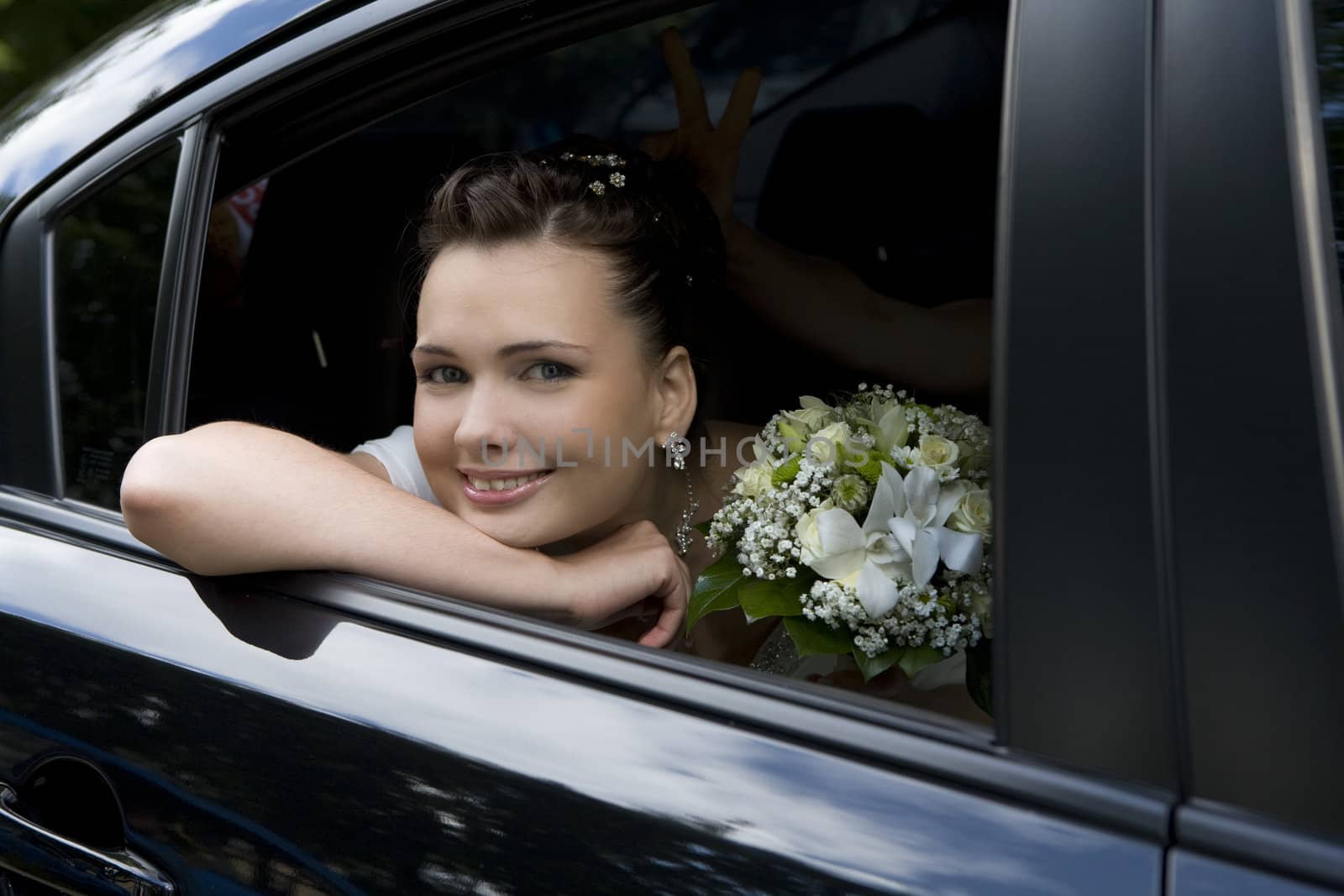 attractive smiling bride with bouquet sitting in wedding car looking through the window
