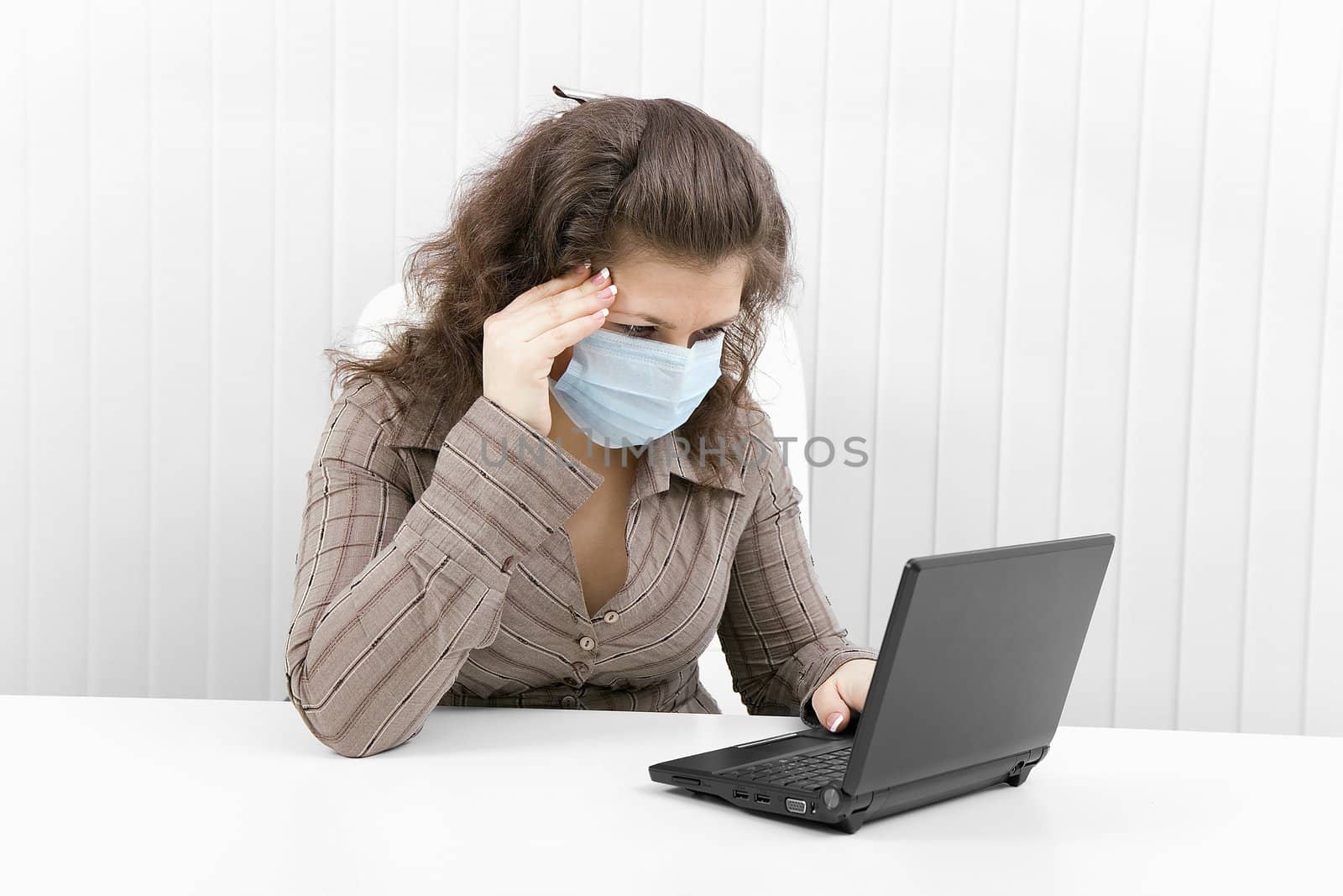 The young woman with medical mask keeps for sick head and works with the laptop