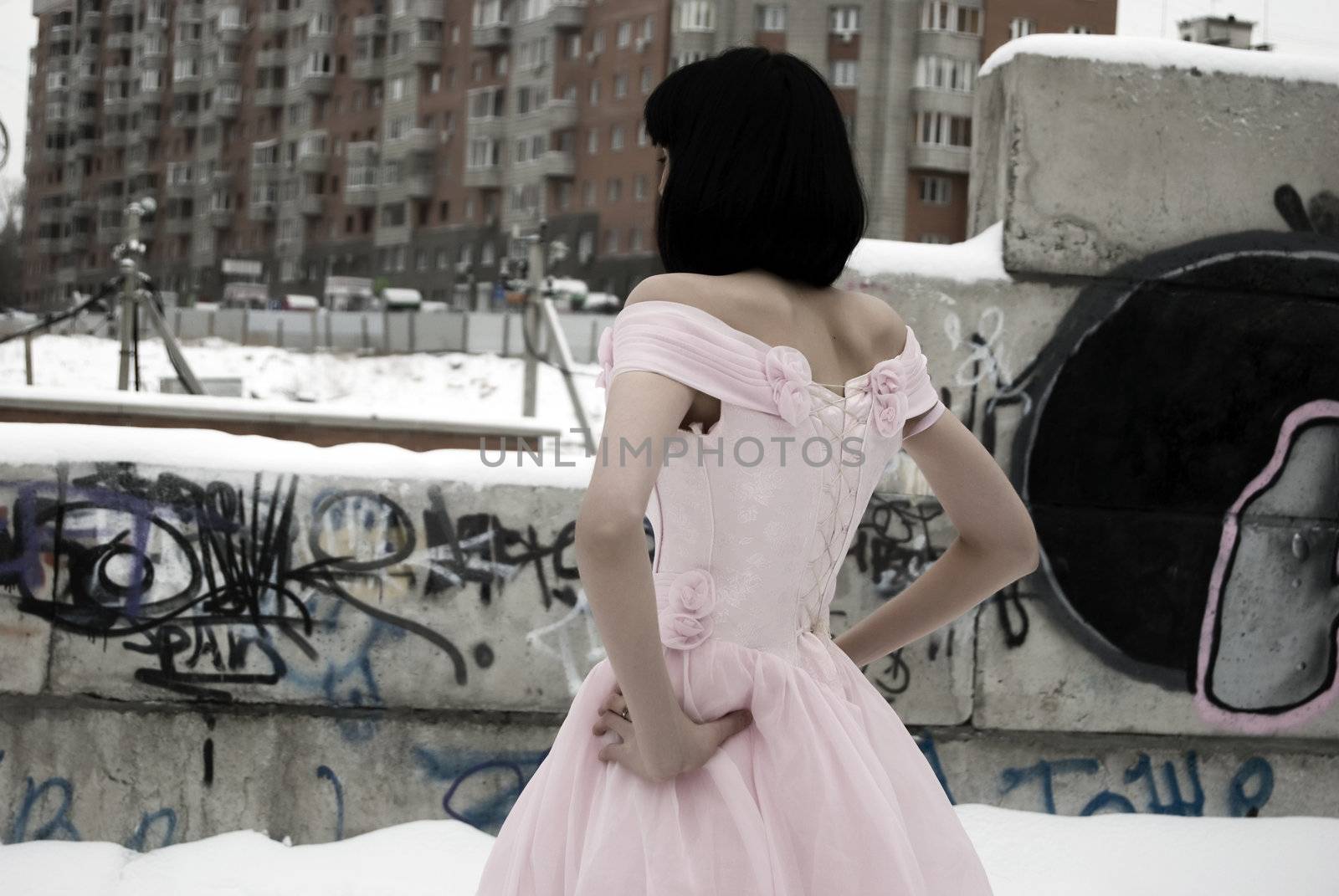 young bride in pink wedding dress standing back to the camera among street ruins 