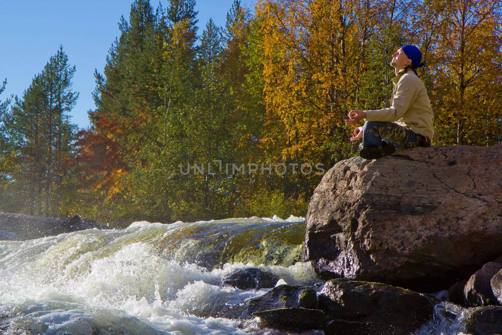 man meditating on a large stone near the waterfall in autumn
