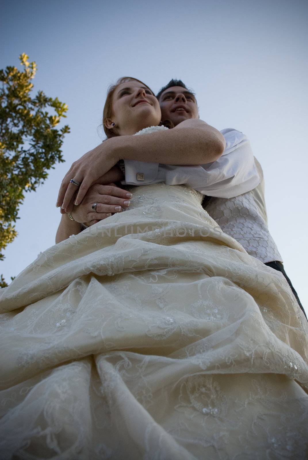 red head sexy beautiful bride in white and groom hugging and standing outside in sun with blue skies