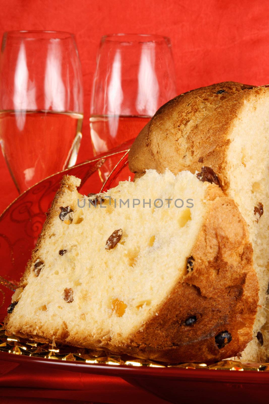 Panettone the italian Christmas fruit cake served on a red glass plate over a red background with golden star decorations, ribbon and two glasses of spumante (sparkling wine). Selective focus.