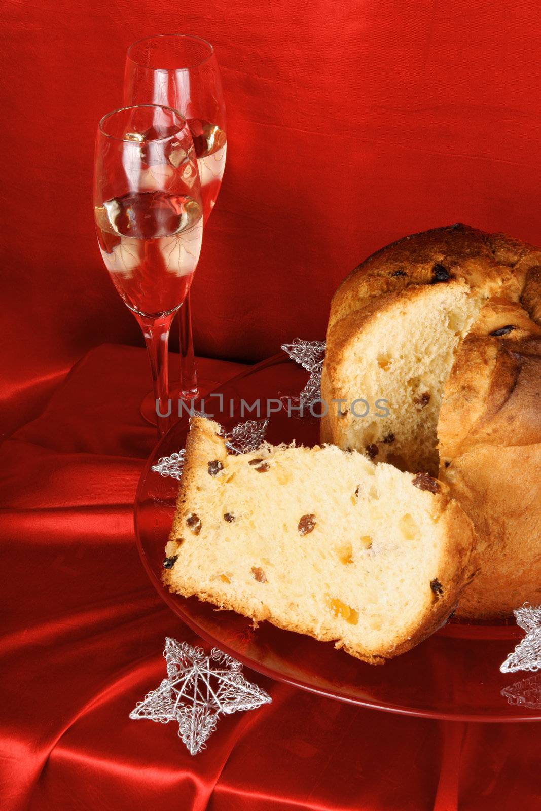 Panettone the italian Christmas fruit cake served on a red glass plate over a red background with silver star decorations and two glasses of sparkling wine (spumante). Selective focus.