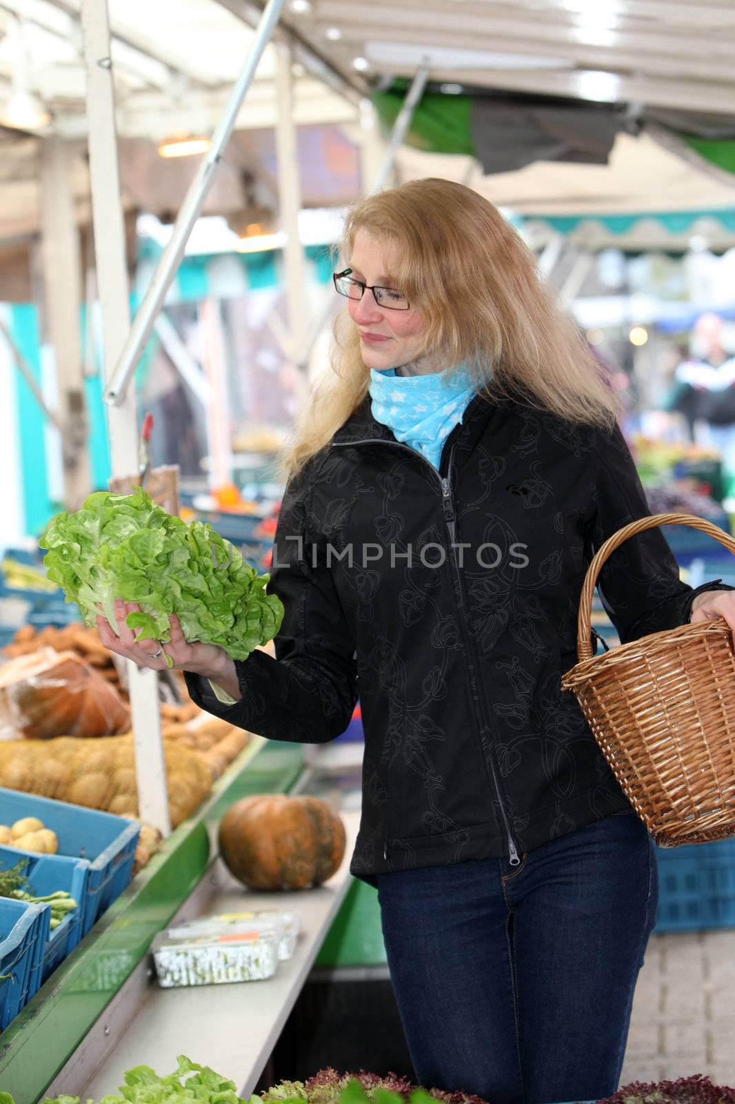 Young woman at a market looking at a head of lettuce and carries a basket
