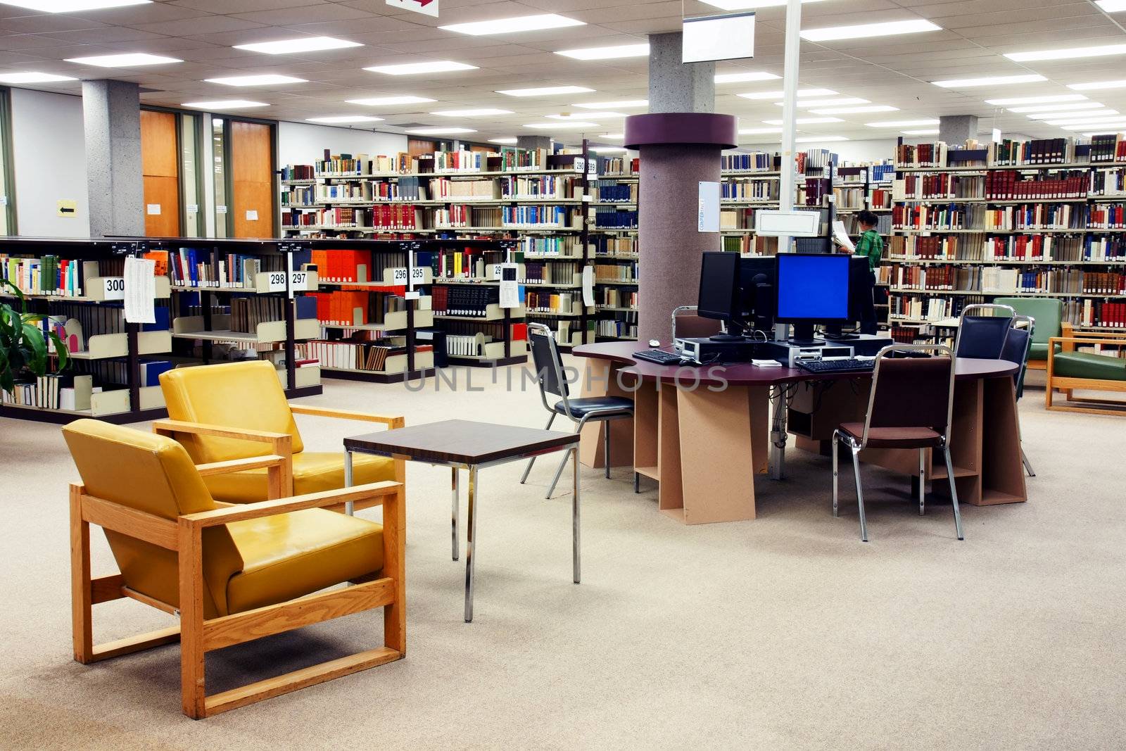 Computer station at the university college library with seating area in the foreground. One young female student searching far in the background.