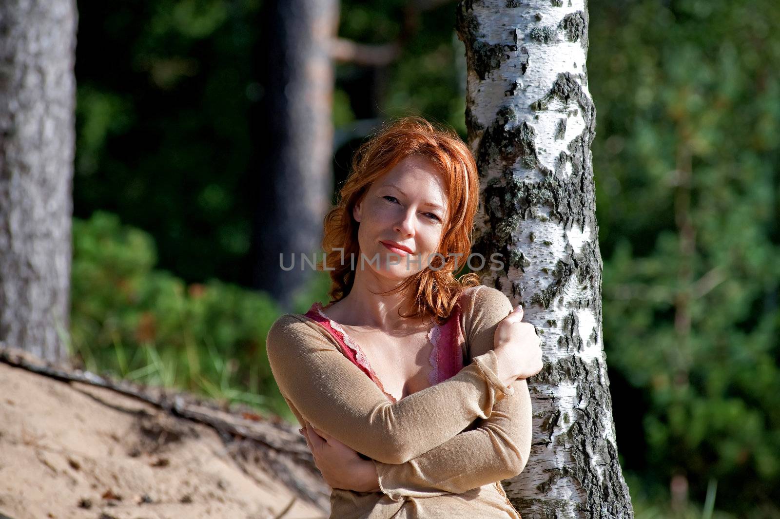 Red-haired adult woman in dune lean on birch.