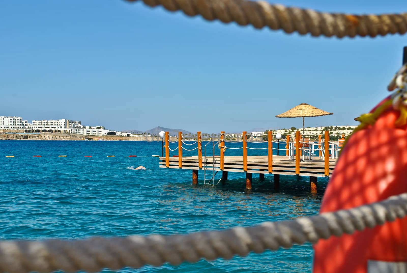 Wooden pontoon seen through two ropes, with blue water and an umbrella