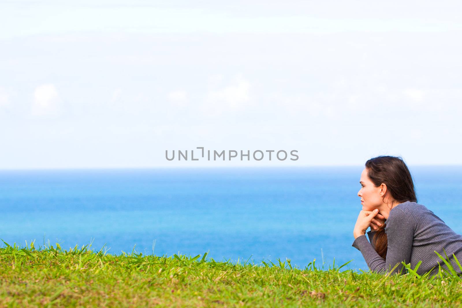 A sad and depressed young woman lying in the green grass looking sadly at the ocean