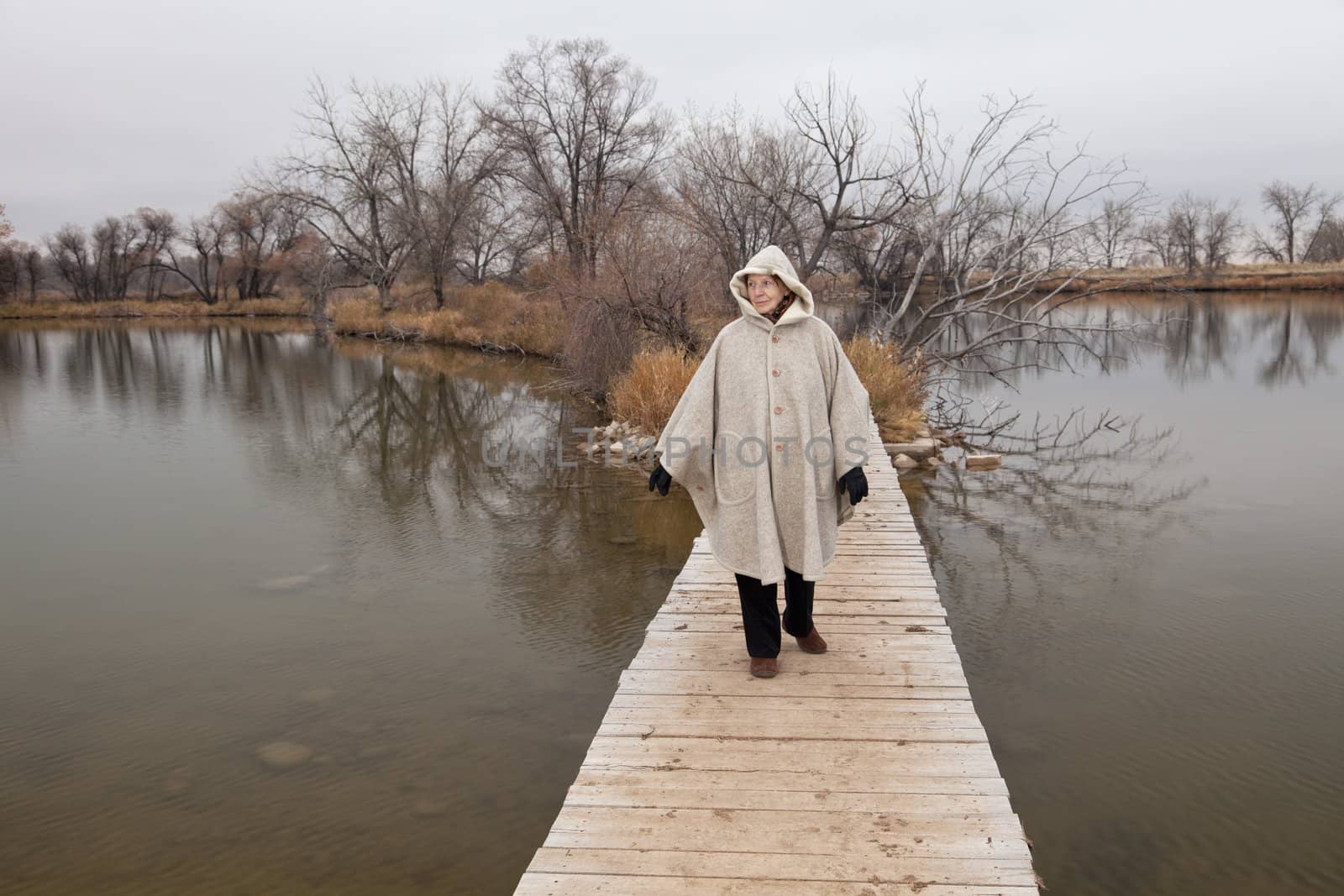 senior woman in her eighties walking alone on a footbridge across lake, nostalgic late fall scenery, life pathway metaphor