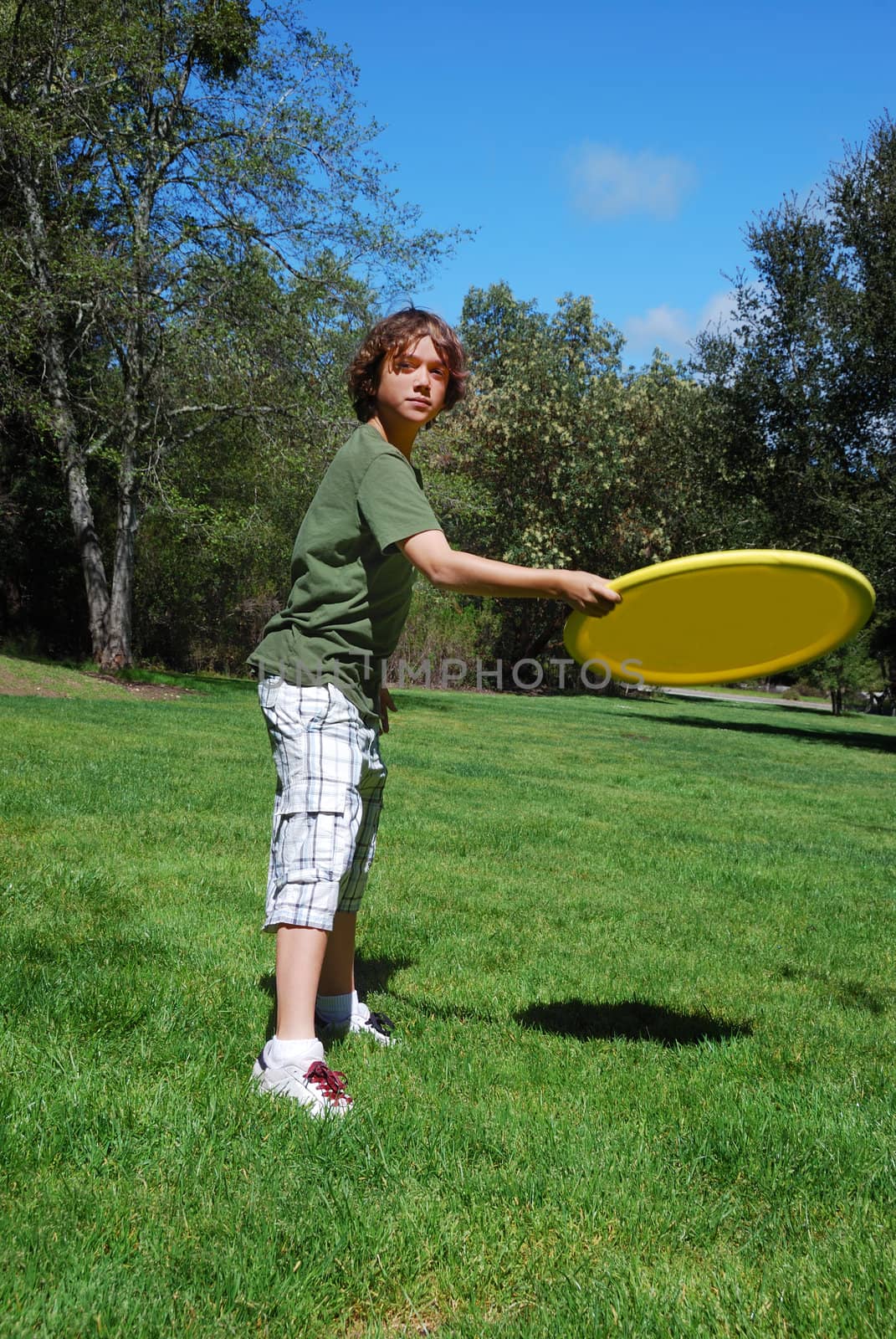 Teen boy throwing yellow frisbee with green grass, trees, and blue sky in the background.