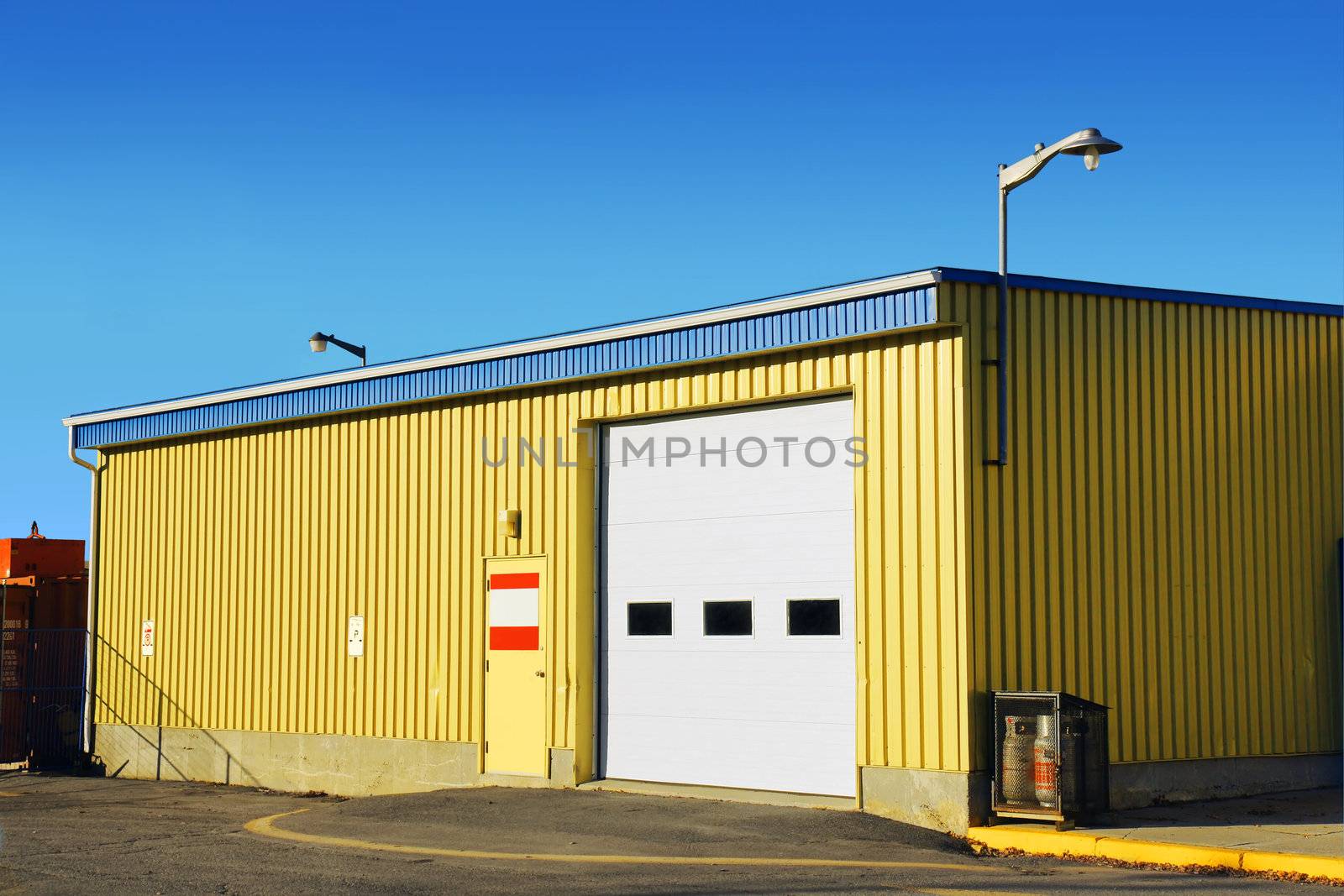 Industrial looking small garage covered with corrugated metal siding, with propane containers on one side and stored crates behind a fence on the other.