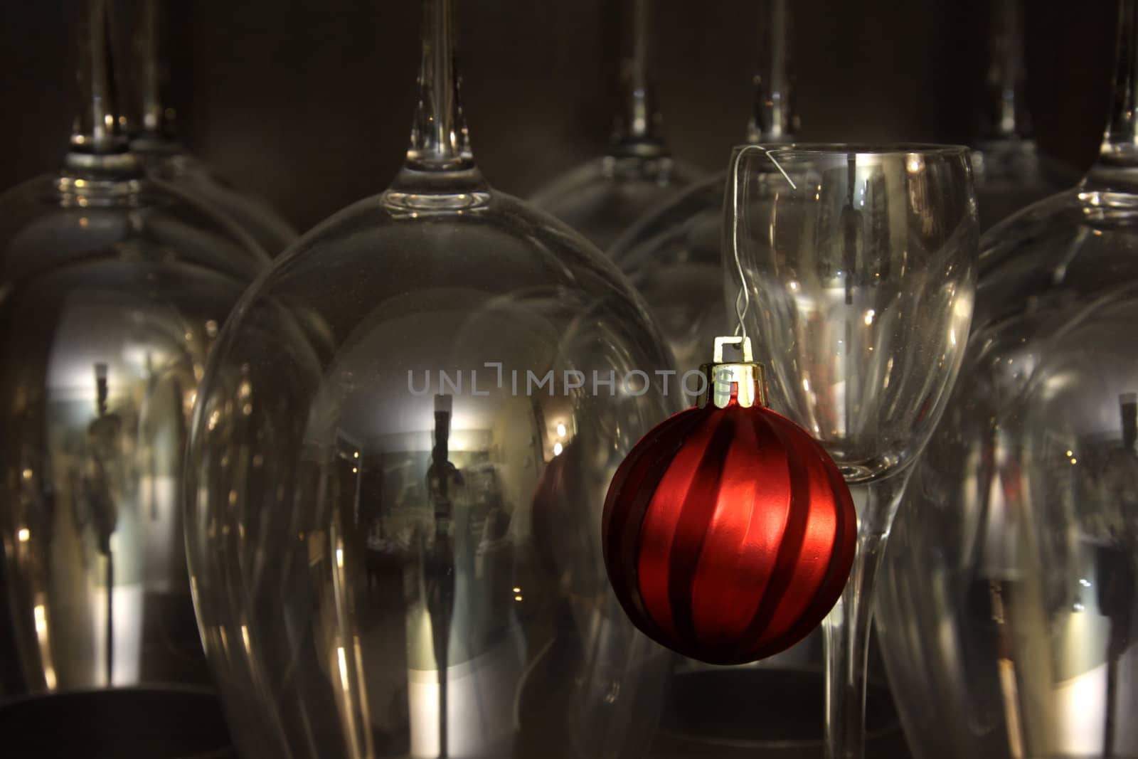 A red Christmas bauble ornament hanging from a dessert glass with wine glasses in the background.
