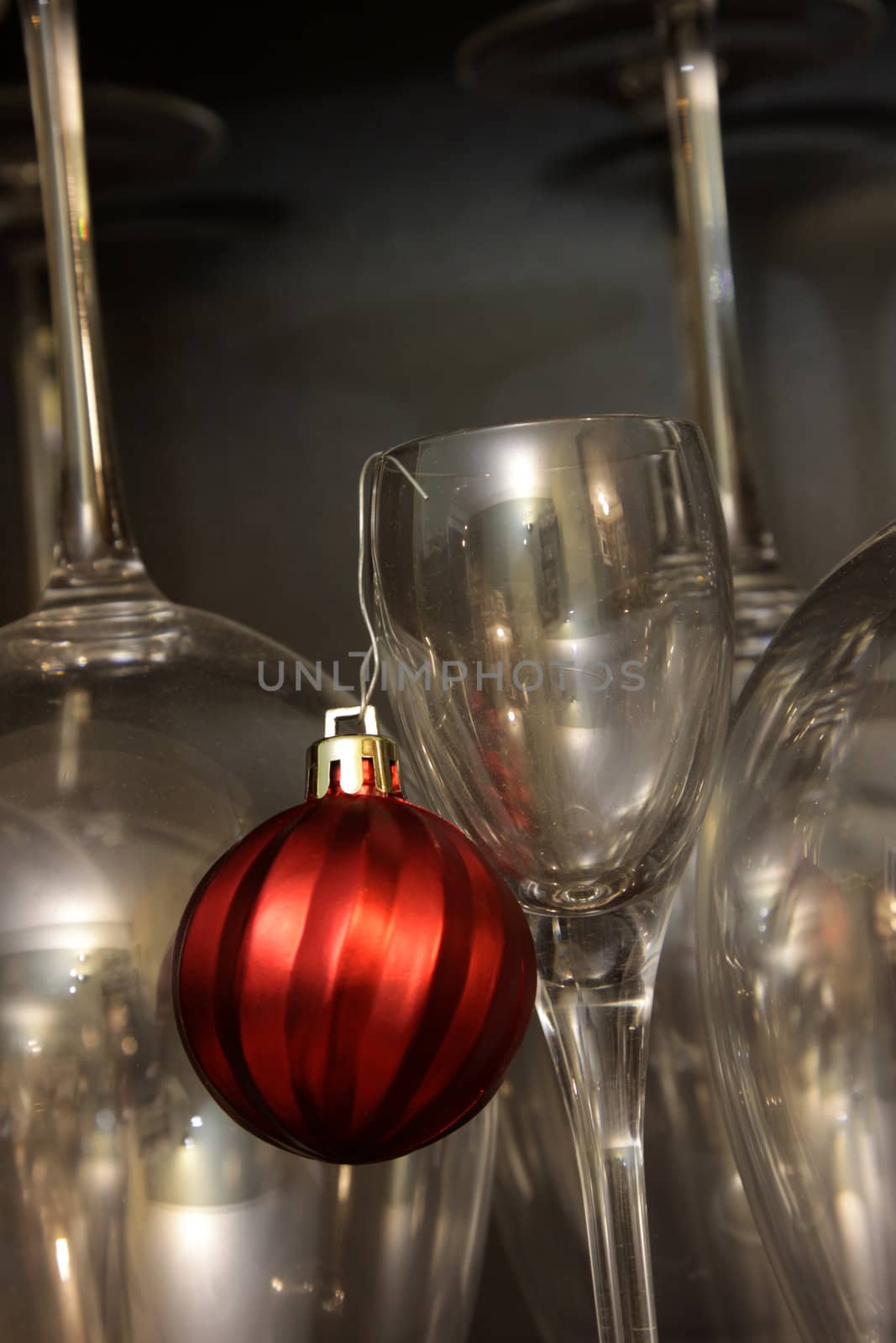A red Christmas bauble ornament hanging from a dessert glass with wine glasses in the background.
