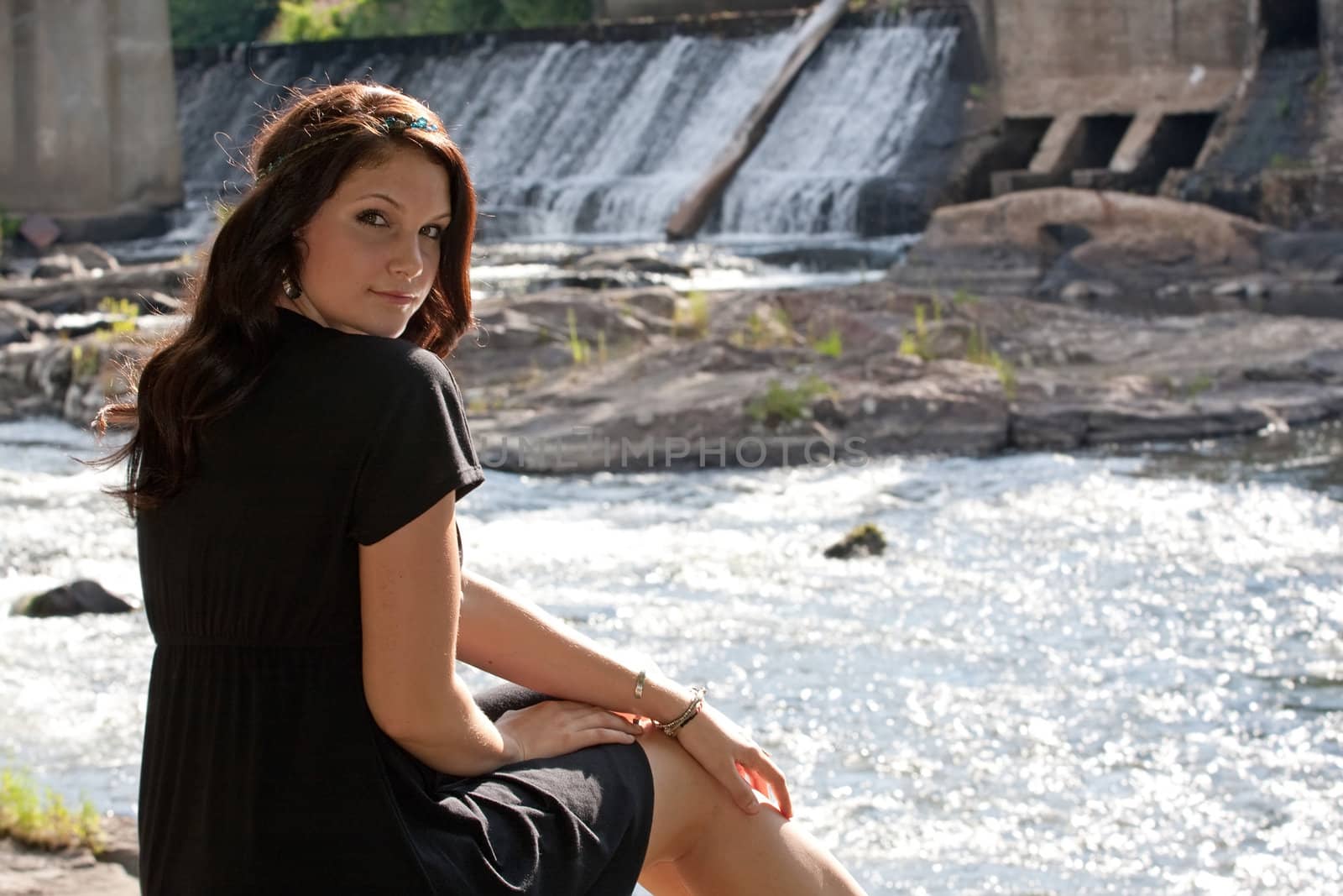 An attractive young woman wearing a black dress sitting by a river.