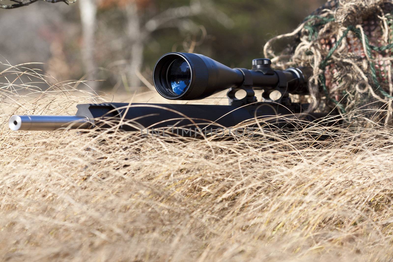 sniper laying on the ground covered in a ghille suite tall grass and trees in the background