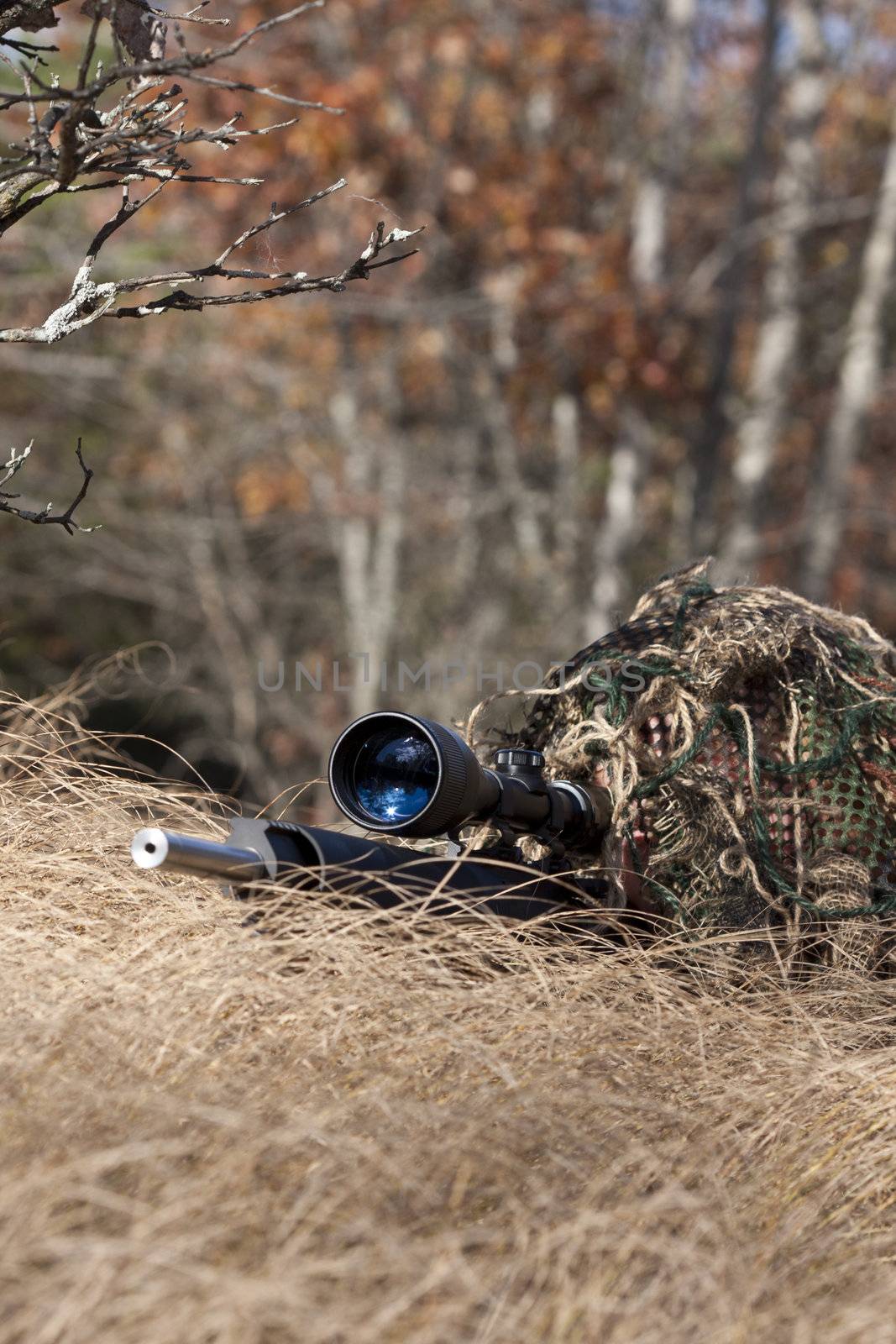 sniper laying on the ground covered in a ghille suite tall grass and trees in the background