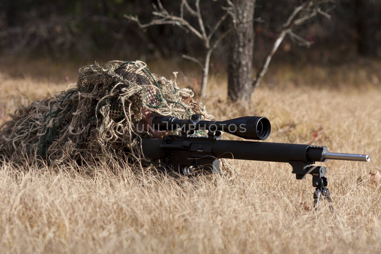 sniper laying on the ground covered in a ghille suite tall grass and trees in the background