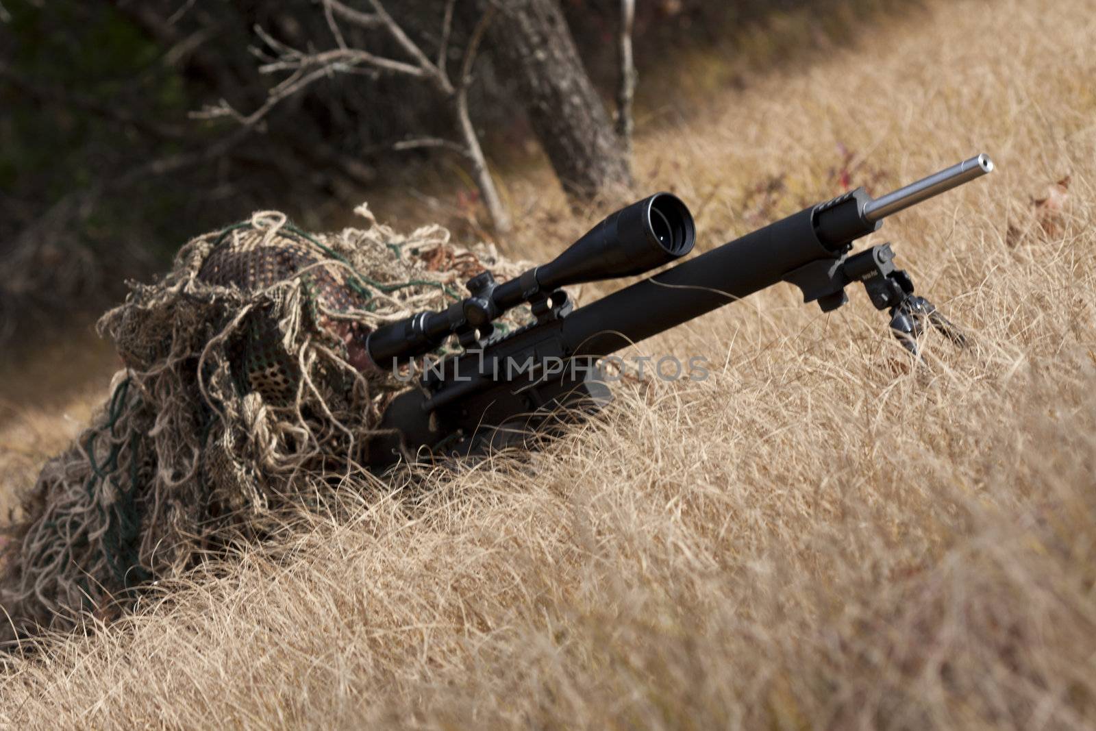 sniper laying on the ground covered in a ghille suite tall grass and trees in the background