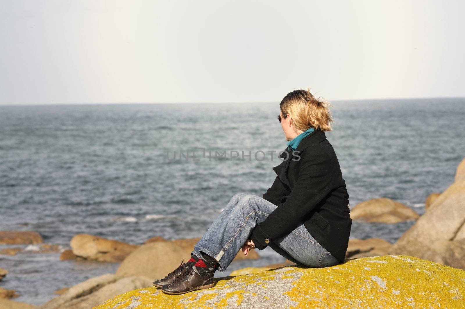 Young woman sitting on granite rock in brittany, watching the atlantic ocean.
