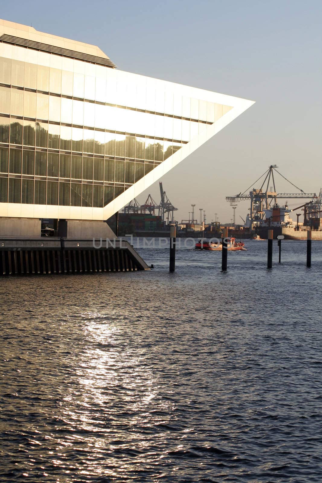 Modern office building 'Dockland' (finished 2005) in Hamburg harbour at sunset. The opposite coastline is reflected in the windows, which again reflect the sunlight onto the water surface. Ships and cranes in the background.