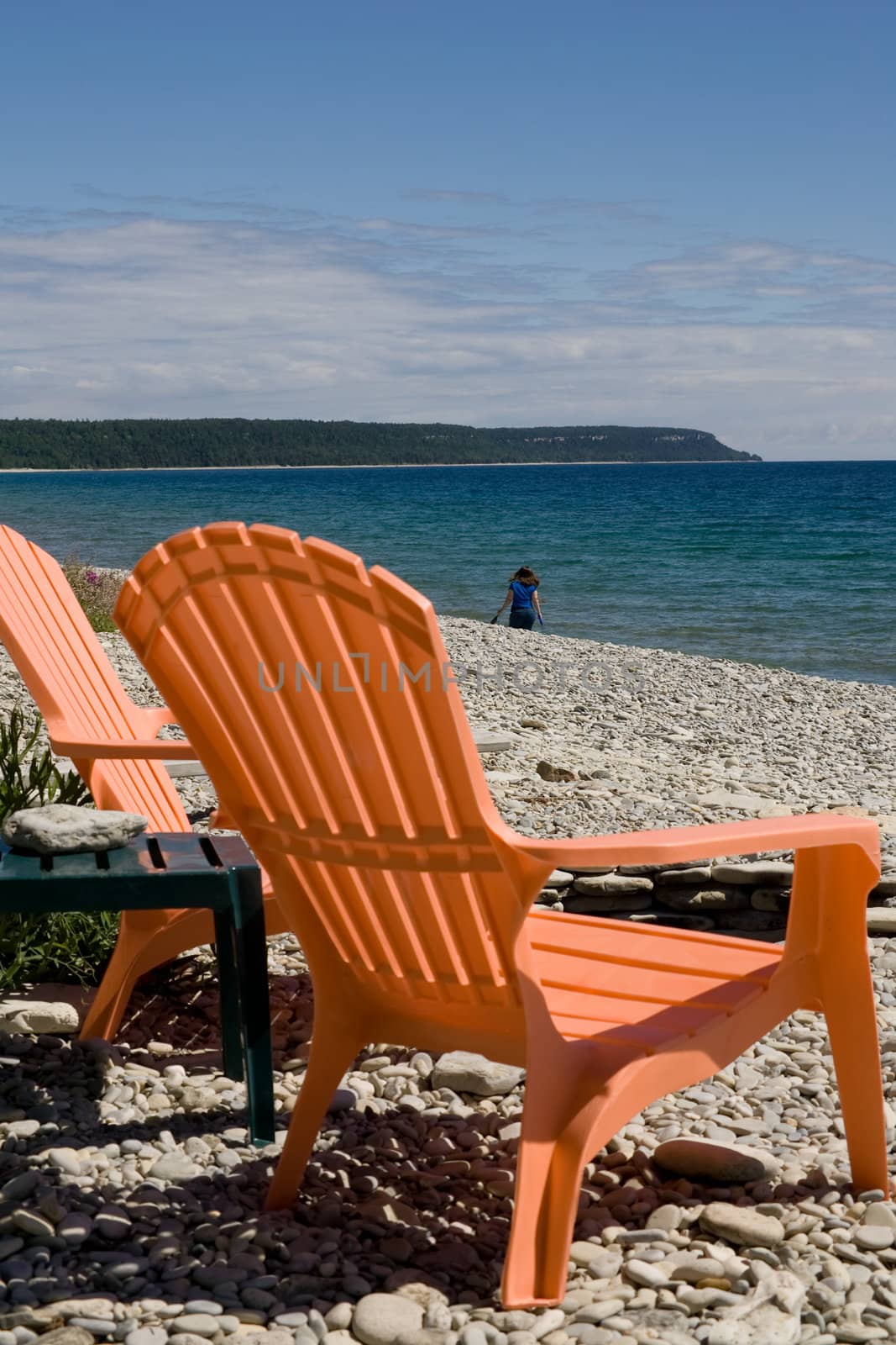 A set of two orange deck chairs with a rocky beach background, low cloud cover, and blue sky.