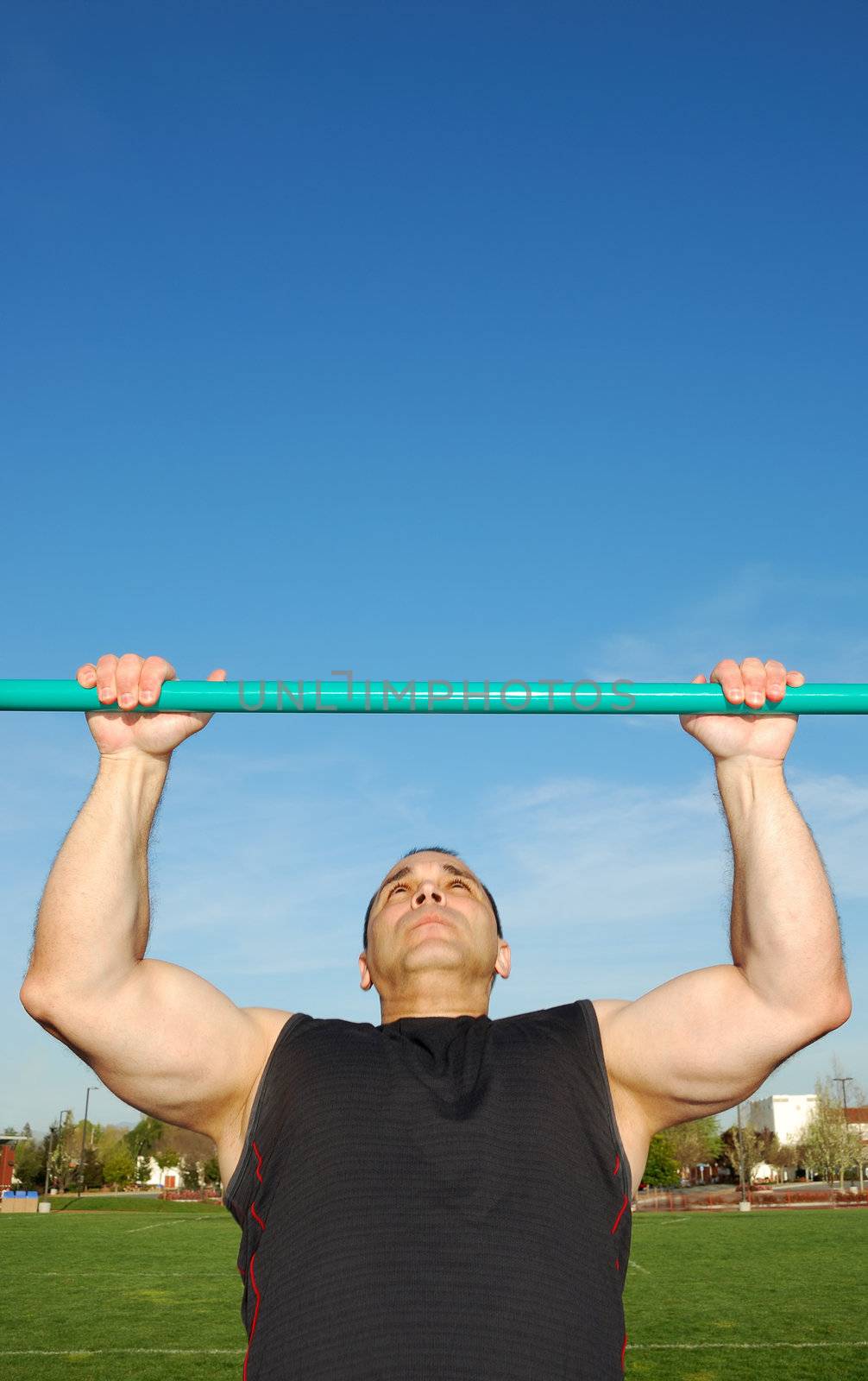 Strong man doing pull ups on a bar in a field with blue sky in the background.