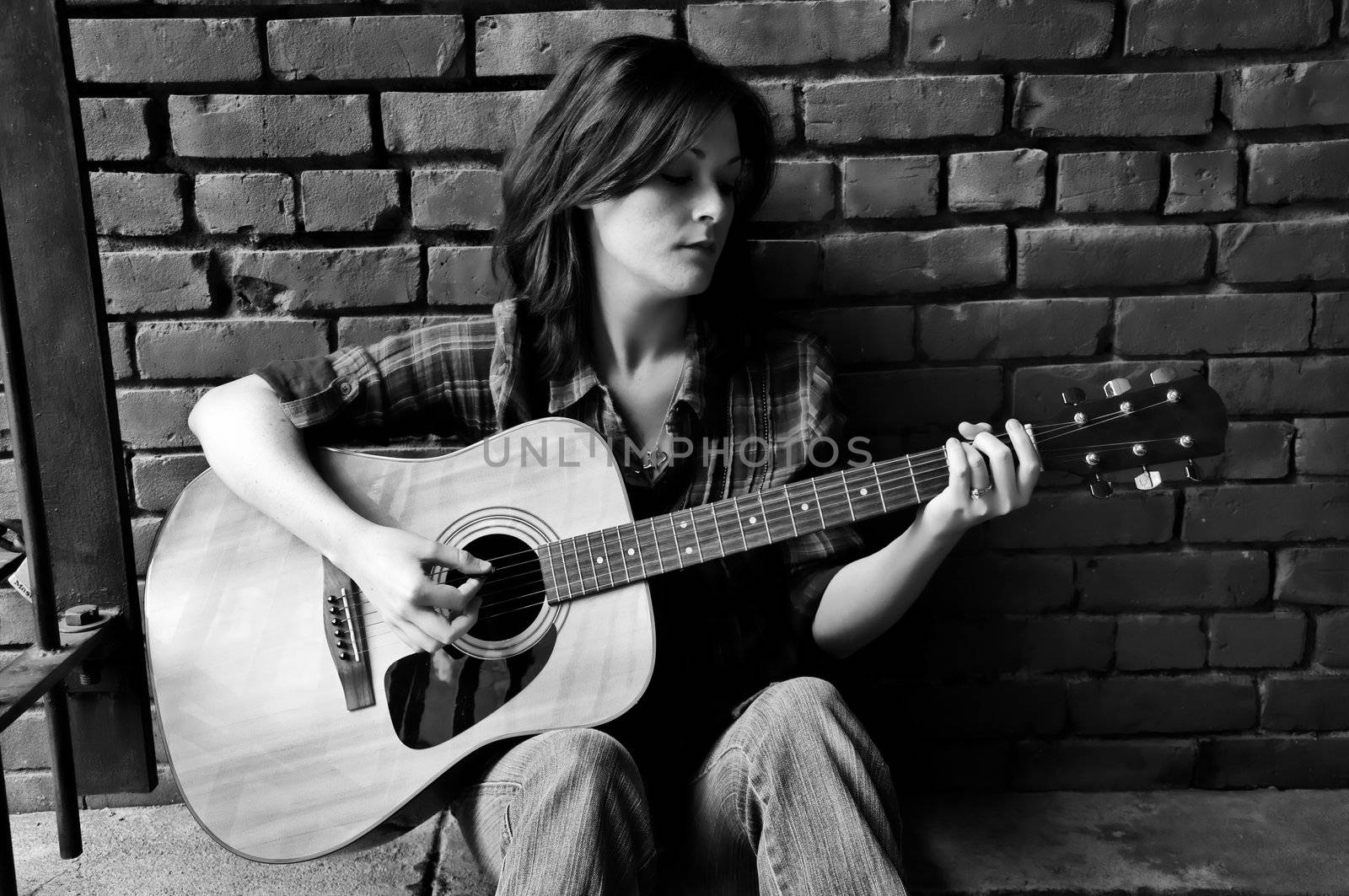 Young female sitting against brick wall and playing guitar.