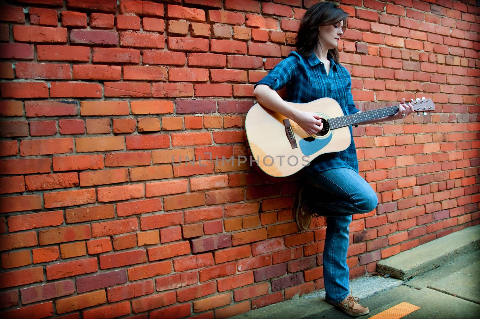 Young female standing in front of  brick wall and playing guitar.