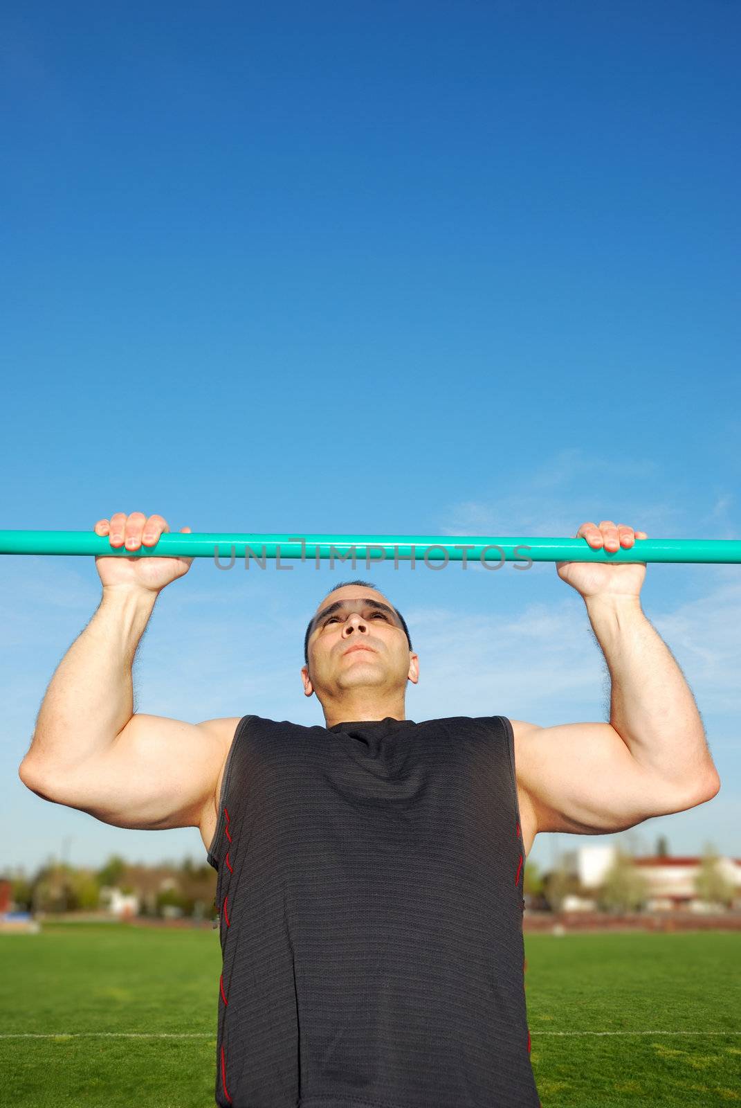 Strong man doing pull ups on a bar in a field with blue sky in the background.