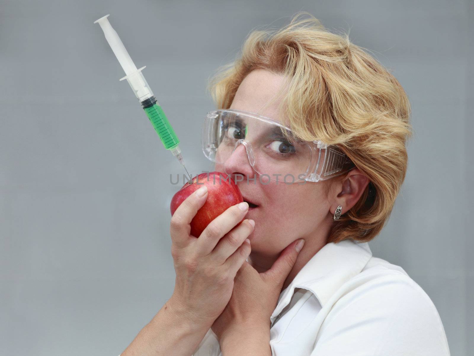 Image of a female researcher trying to eat an apple with a syringe with solution in it. Useful image to suggest the danger of eating genetically transformed food or to promote pure natural food.
