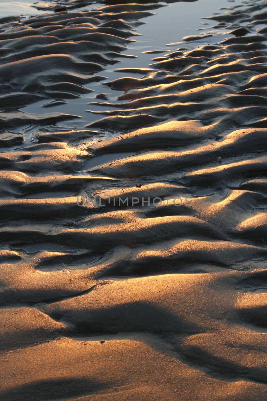 Sand textured with abstract patterns of waves, lines and ruts highlighted by red light and shadows