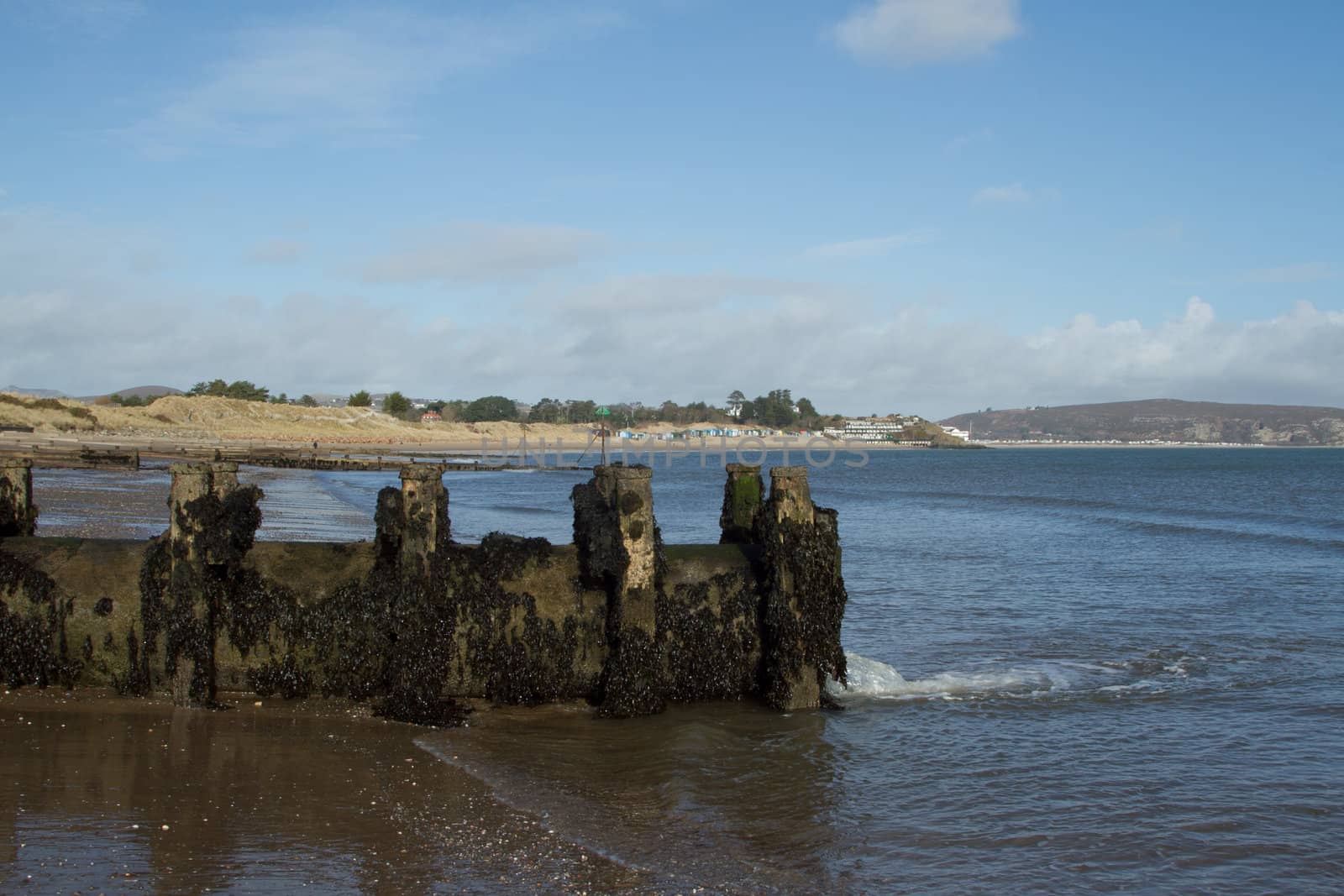 Water drainage pipe made of metal with wooden supports on a beach, water is flowing into the sea, a popular beach destination in the distance.