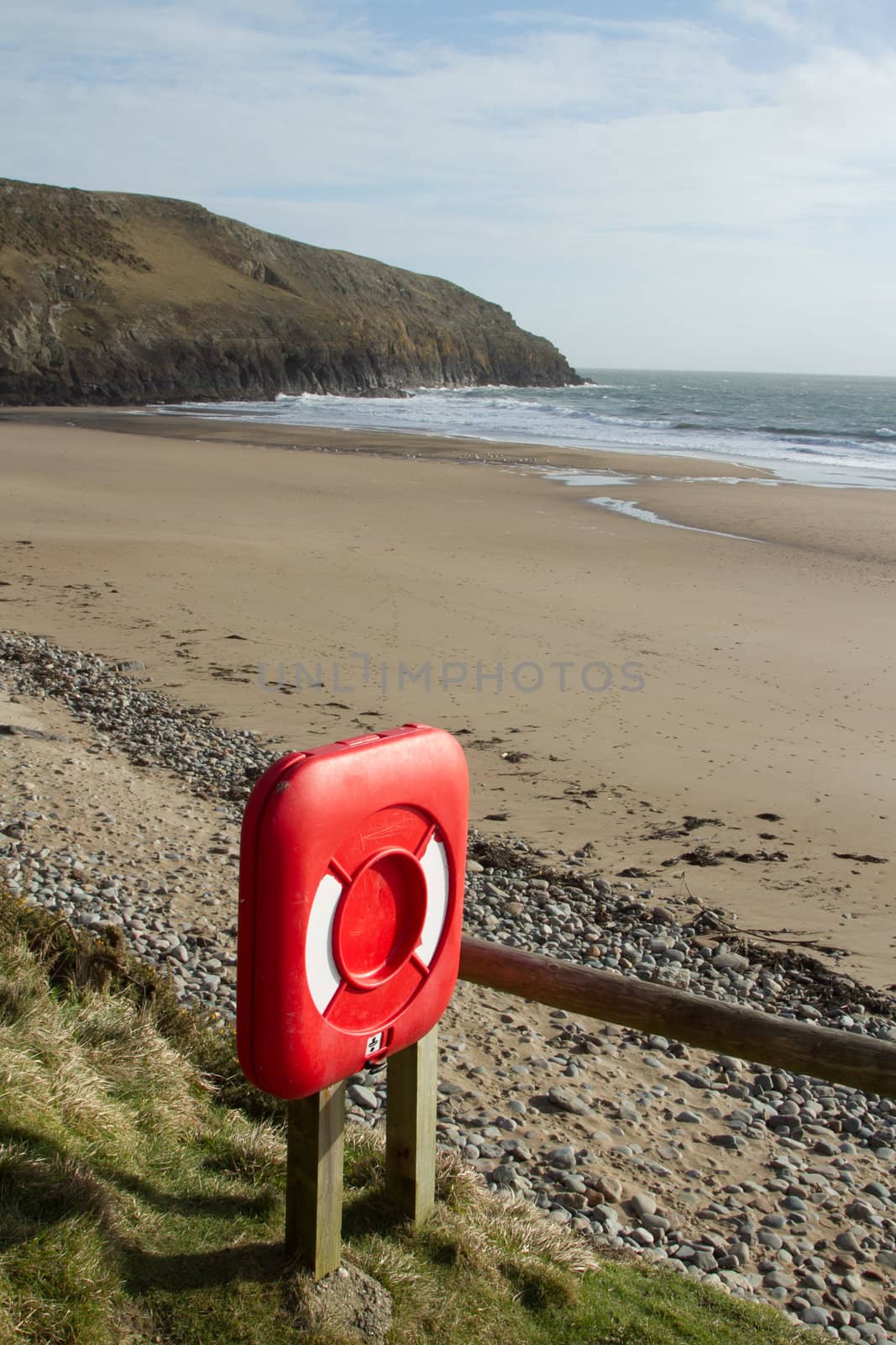 A red container, life saving equipment, placed by a beach with sand, sea and cliff.