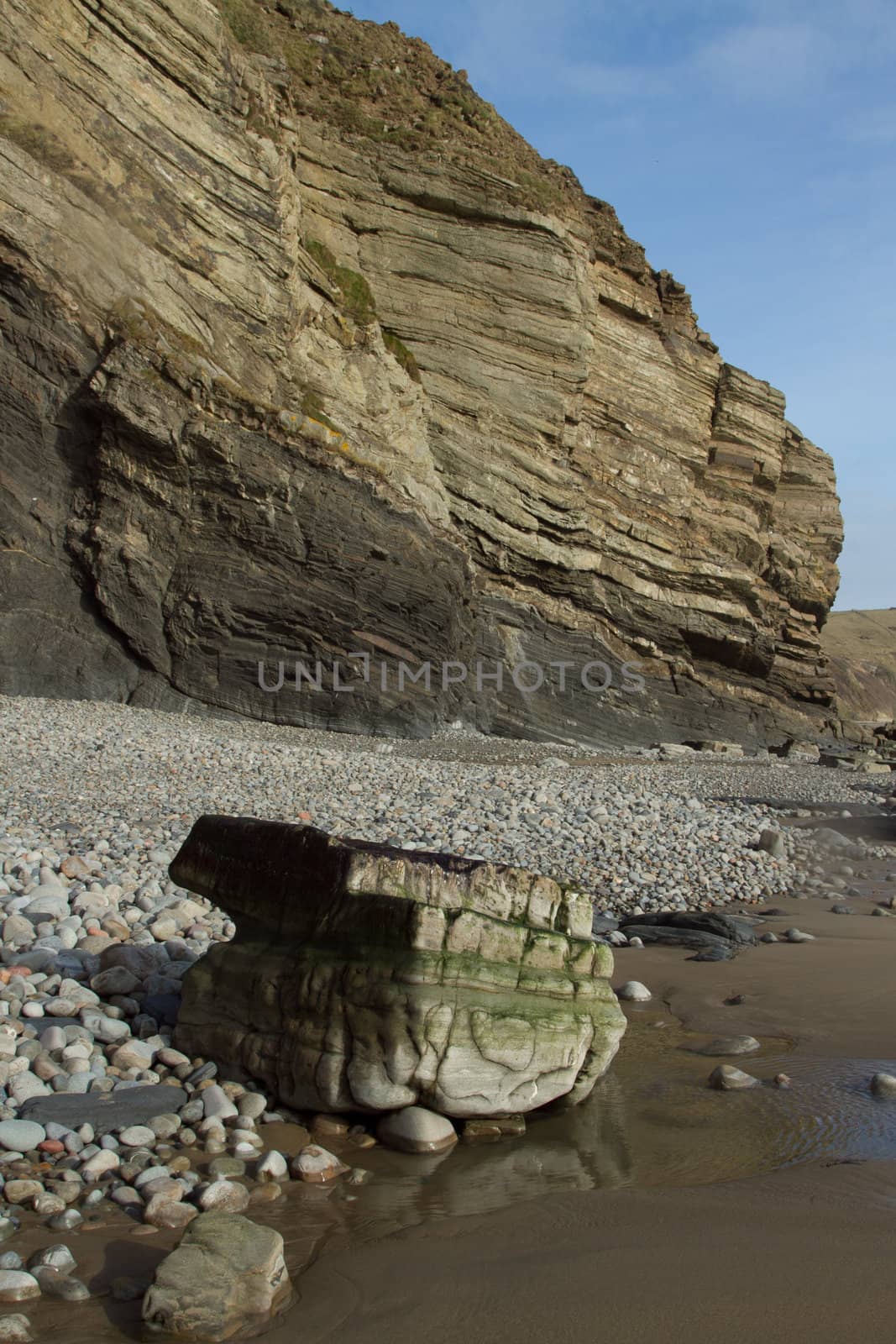 Geological feature, striated shale and mud-stone cliff on a beach with pebble and a water smoothed boulder on the sand.