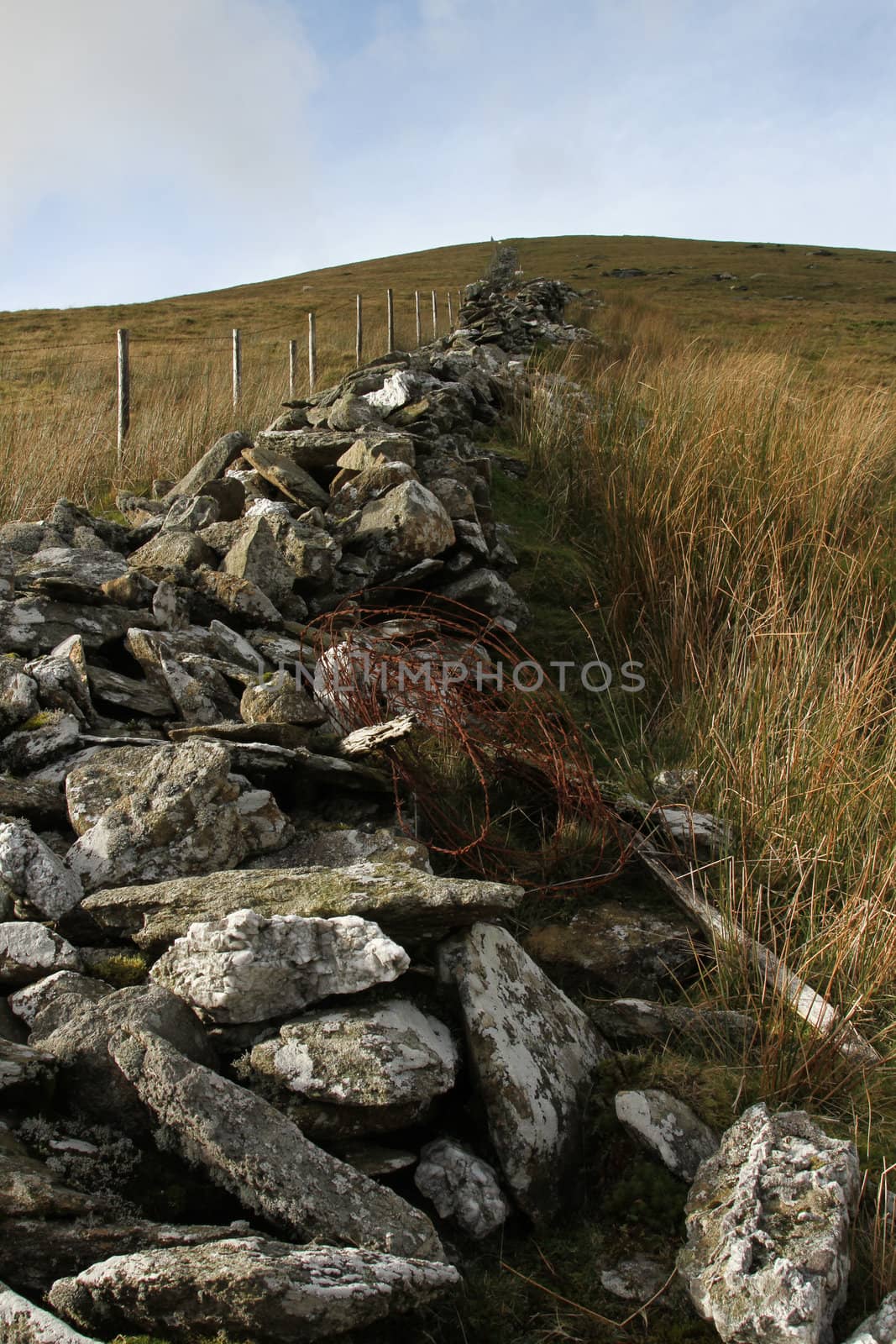 Tumble down stone wall with rusty barbed wire and a new fence adjacent with posts running up a hillside.