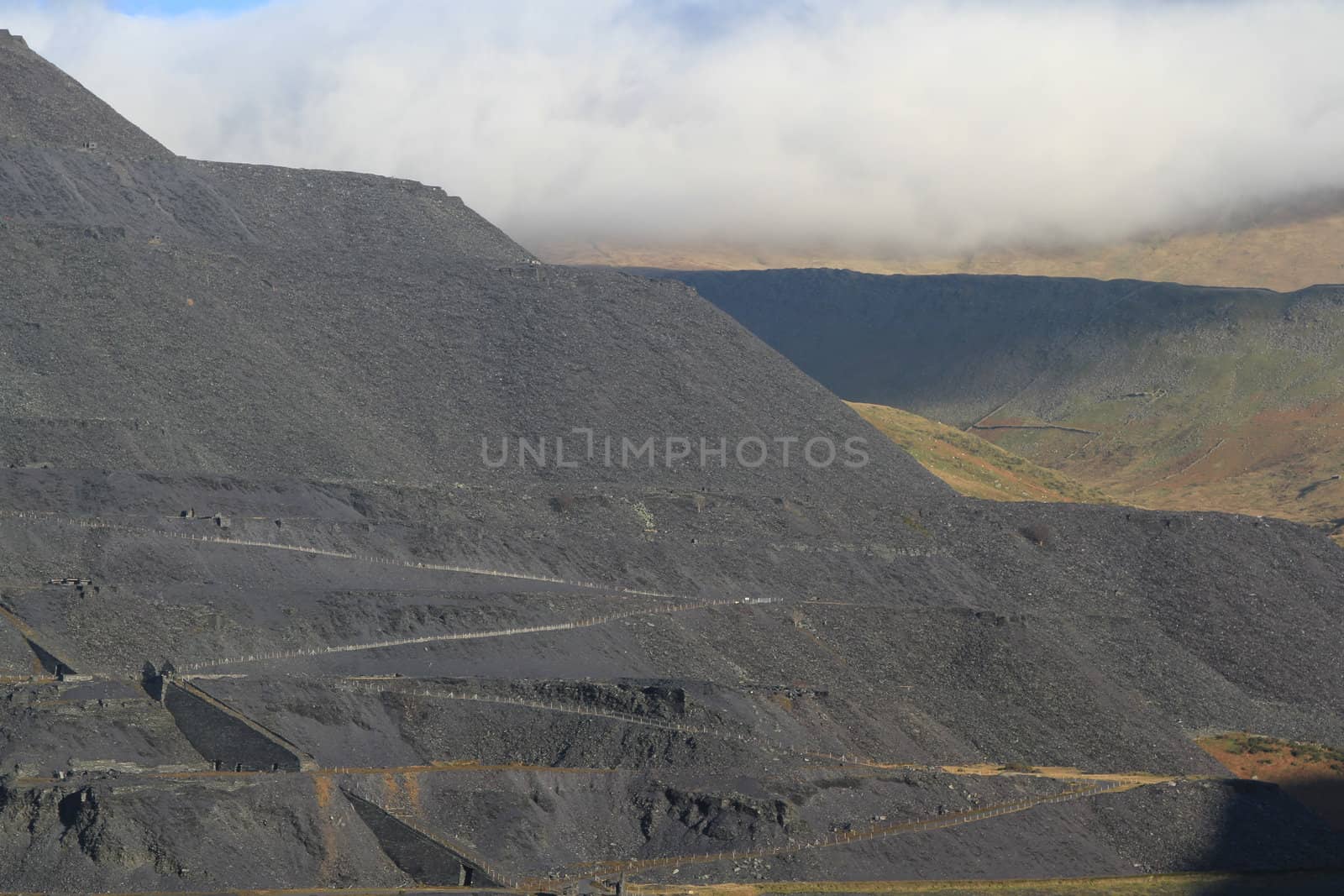 Waste products of slate quarrying in levels on a mountainside with zigzag paths and fences.