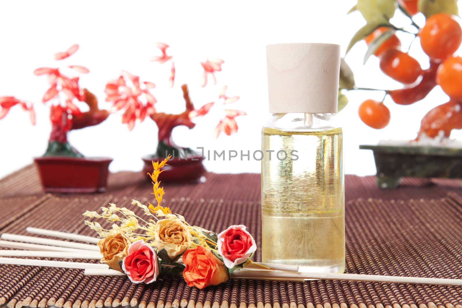 Glass bottle with yellow oil and small flowers lay on brown mat. Stone trees at the background
