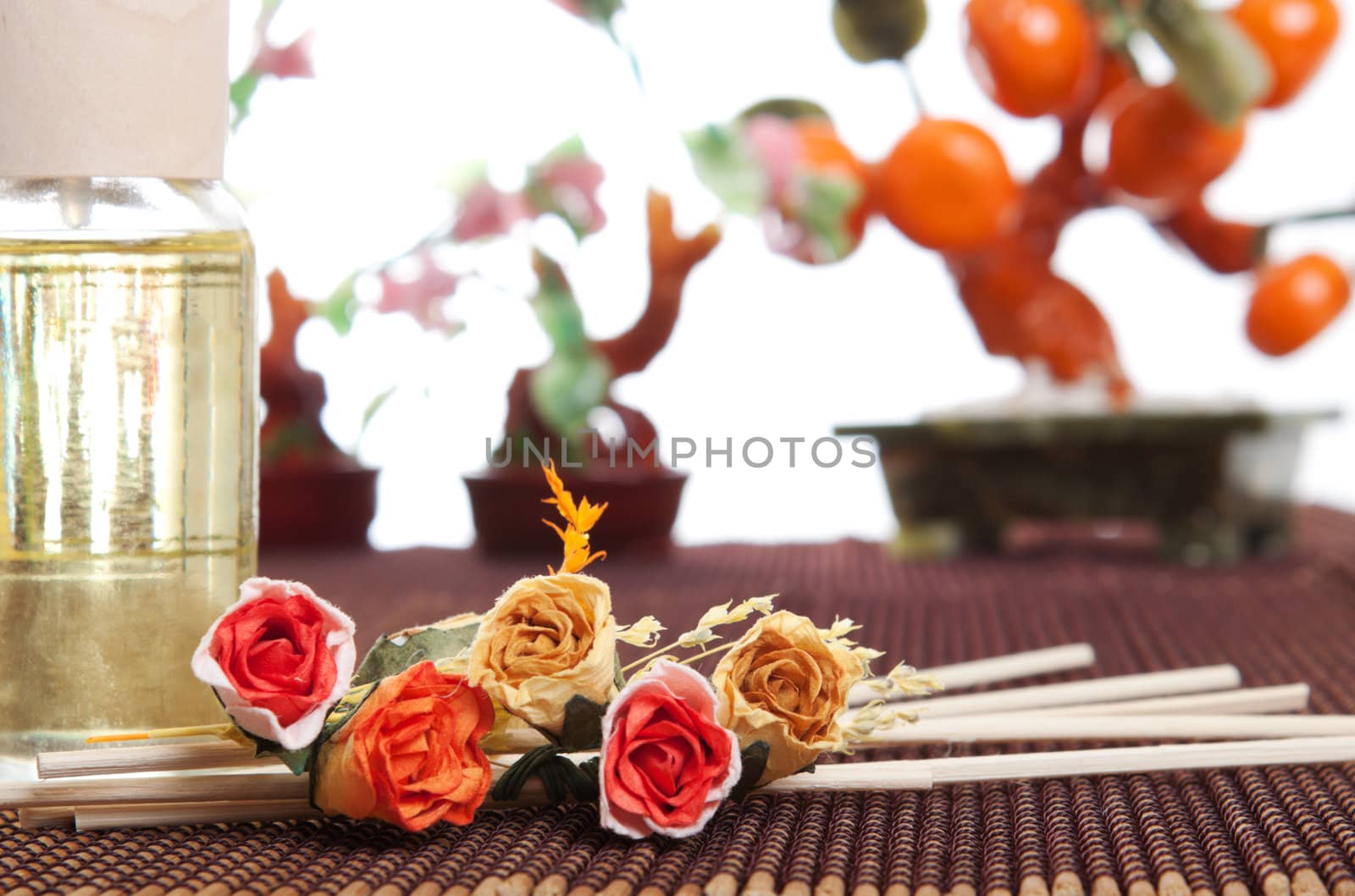 Glass bottle with yellow oil and small flowers lay on brown mat. Stone trees at the background