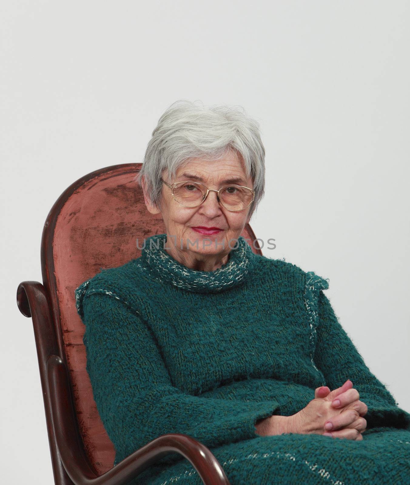 Portrait of an old woman sitting on a wooden rocking chair with fingers crossed, against a grey background.