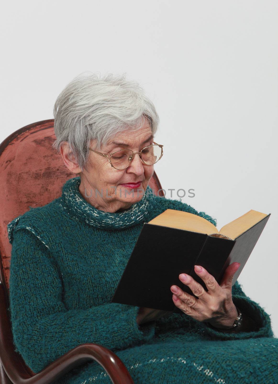 Portrait of an old woman reading a black book against a grey background.