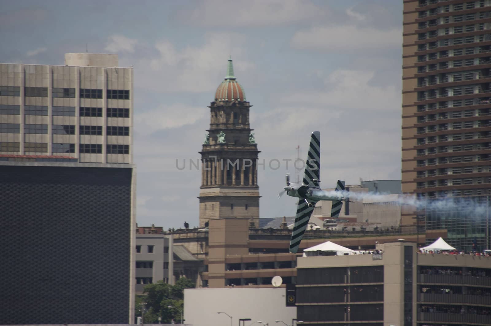 Air race plane doing stunt flying through the city with a crowd of people watching in the background.