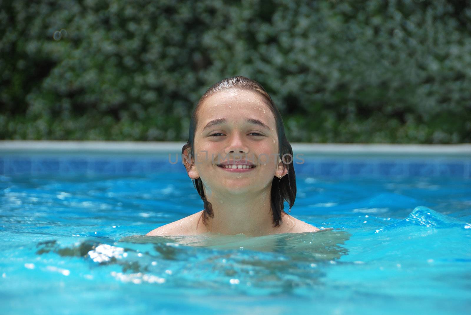 Smiling teen boy in swimming pool surrounded with white flower bushes in the background.