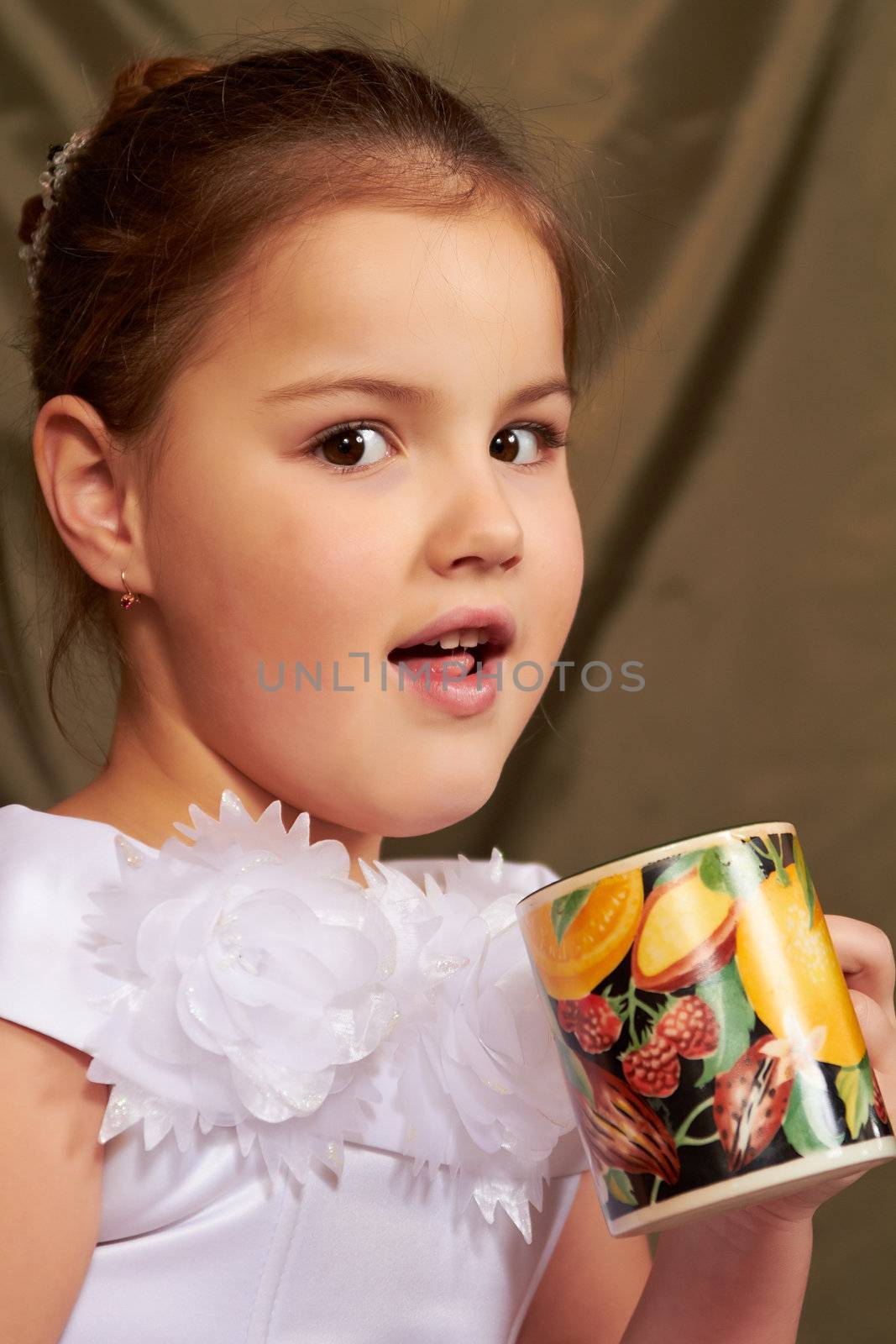 The little girl in a white dress drinks from a large mug.