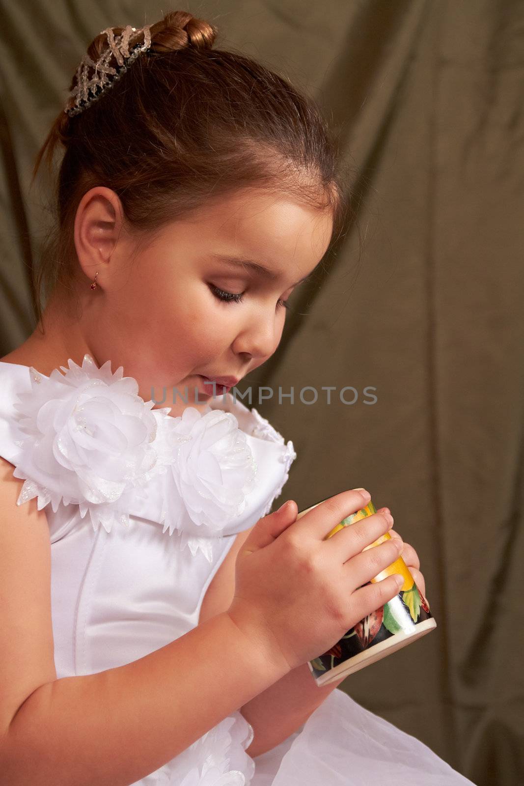 The little girl in a white dress drinks from a large mug.