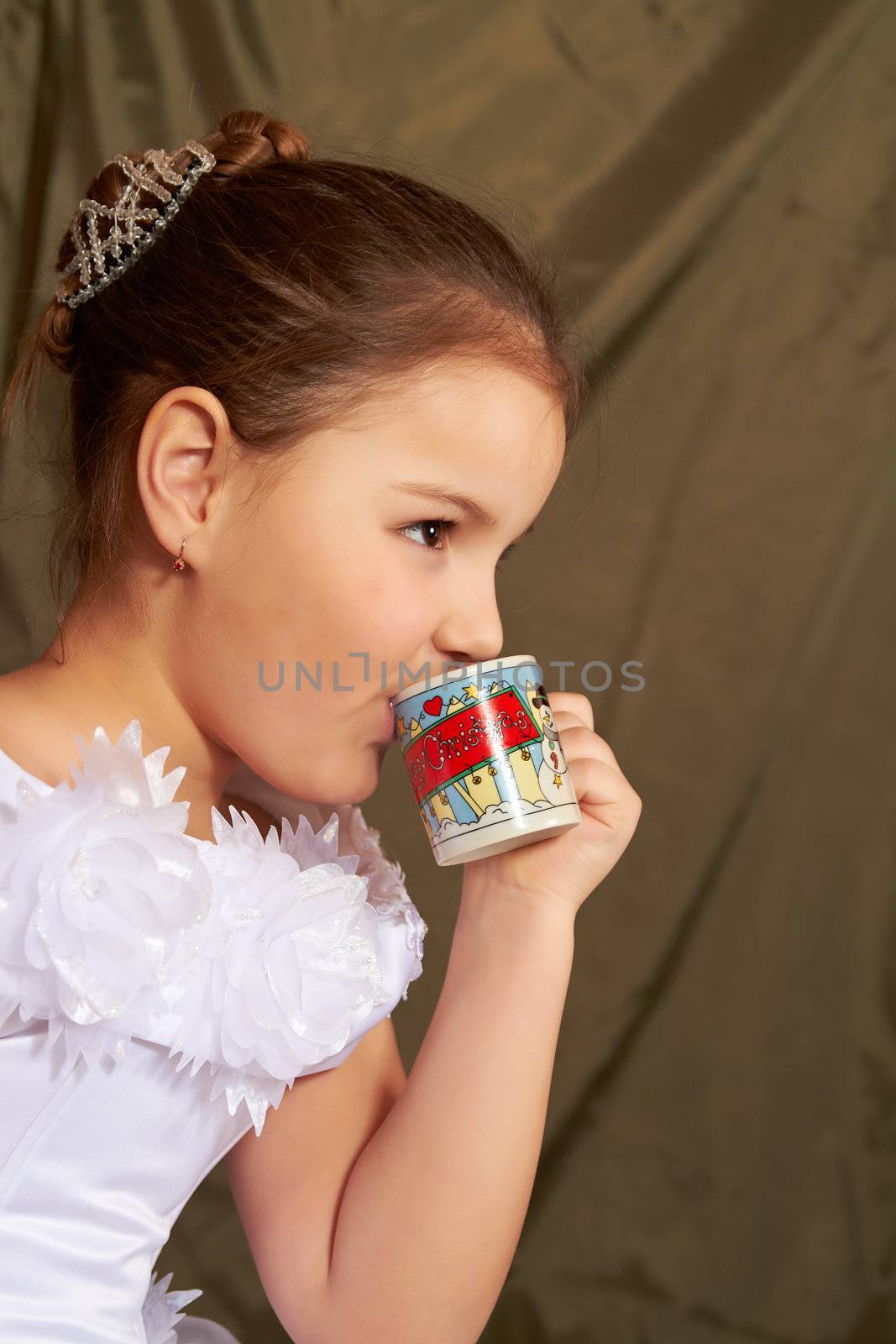 The little girl in a white dress drinks from a small cup.