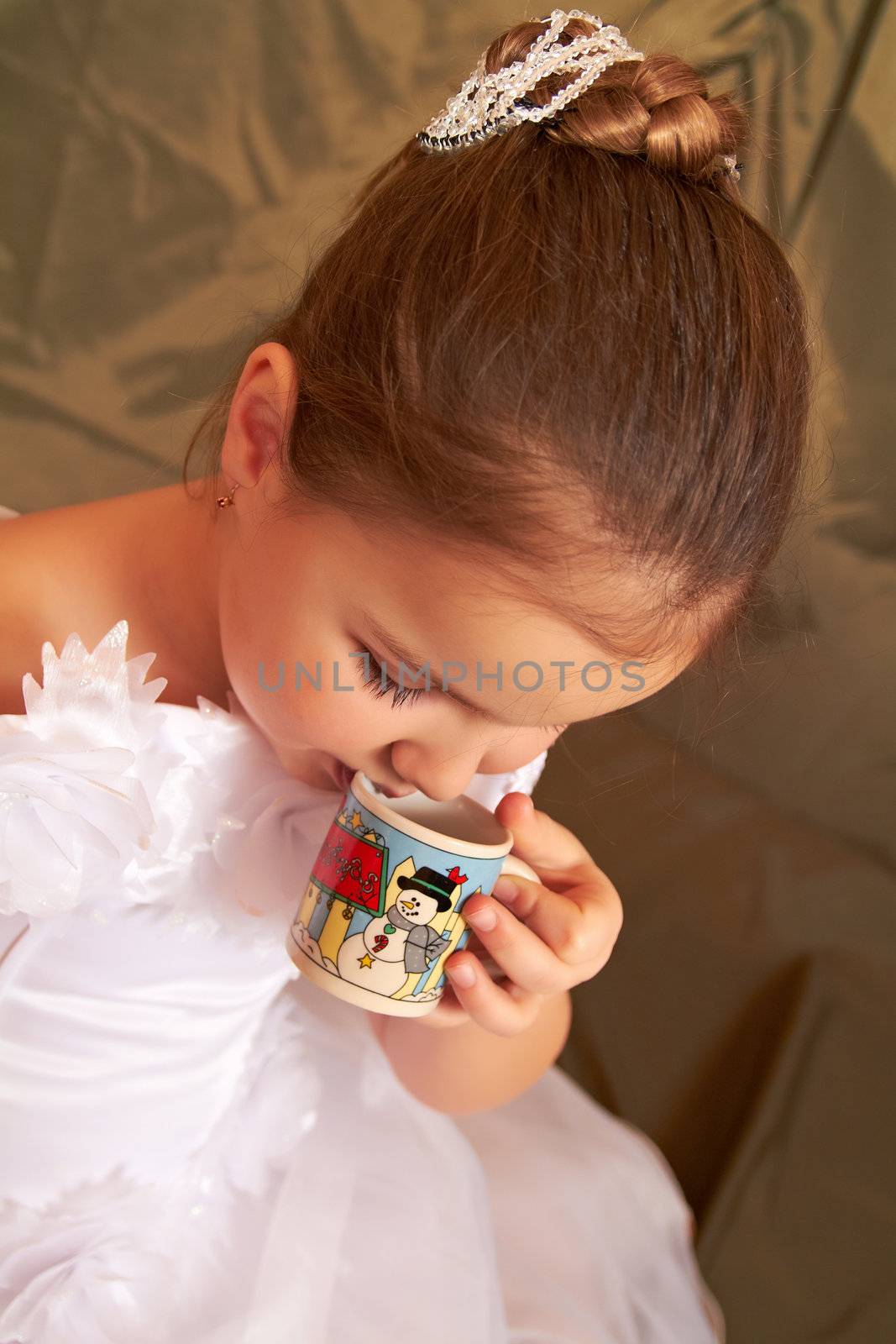 The little girl in a white dress drinks from a small cup.