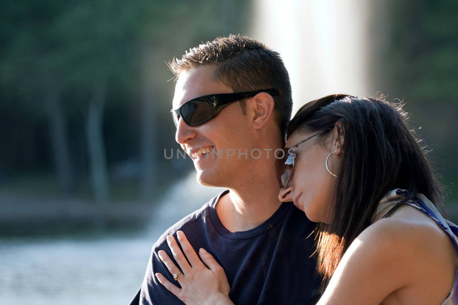 A happy young couple in their mid 20s wearing sunglasses with the girl leaning on the guys shoulder.